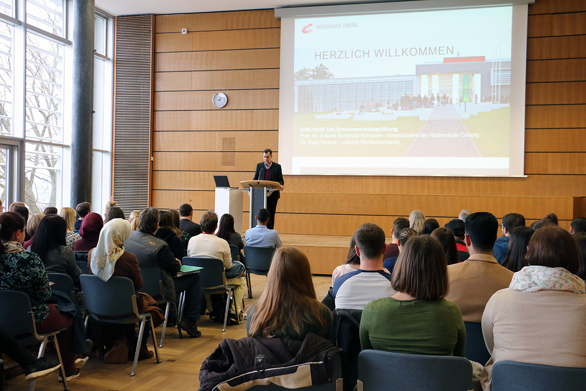 Eine Person steht an einem Rednerpult und hält einen Vortrag vor einem Publikum in einem Hörsaal der Hochschule Coburg. Auf einem großen Bildschirm werden ein Begrüßungstext und ein Bild des modernen Campusgebäudes angezeigt. Die Teilnehmer blicken nach vorne und hören aufmerksam zu.