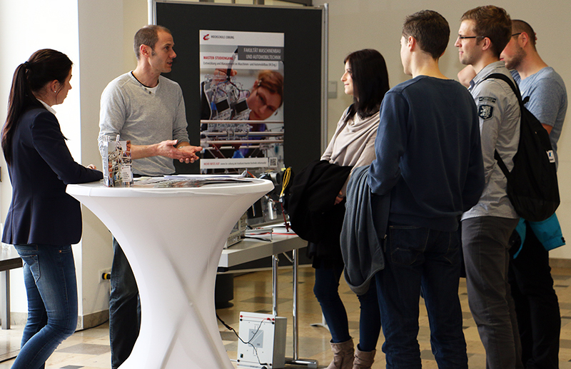 Eine Gruppe von Menschen steht bei einer Technologieausstellung der Hochschule Coburg um einen großen weißen Tisch herum. Ein Mann präsentiert mehreren Besuchern Informationen, während im Hintergrund Poster mit technologiebezogenen Grafiken zu sehen sind.