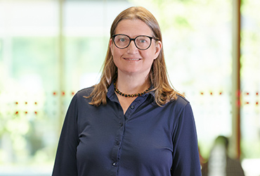 A person with long hair and glasses is smiling at the camera, wearing a Hochschule Coburg button-up shirt and a necklace. The background is blurred and bright, suggesting an indoor setting with natural light.