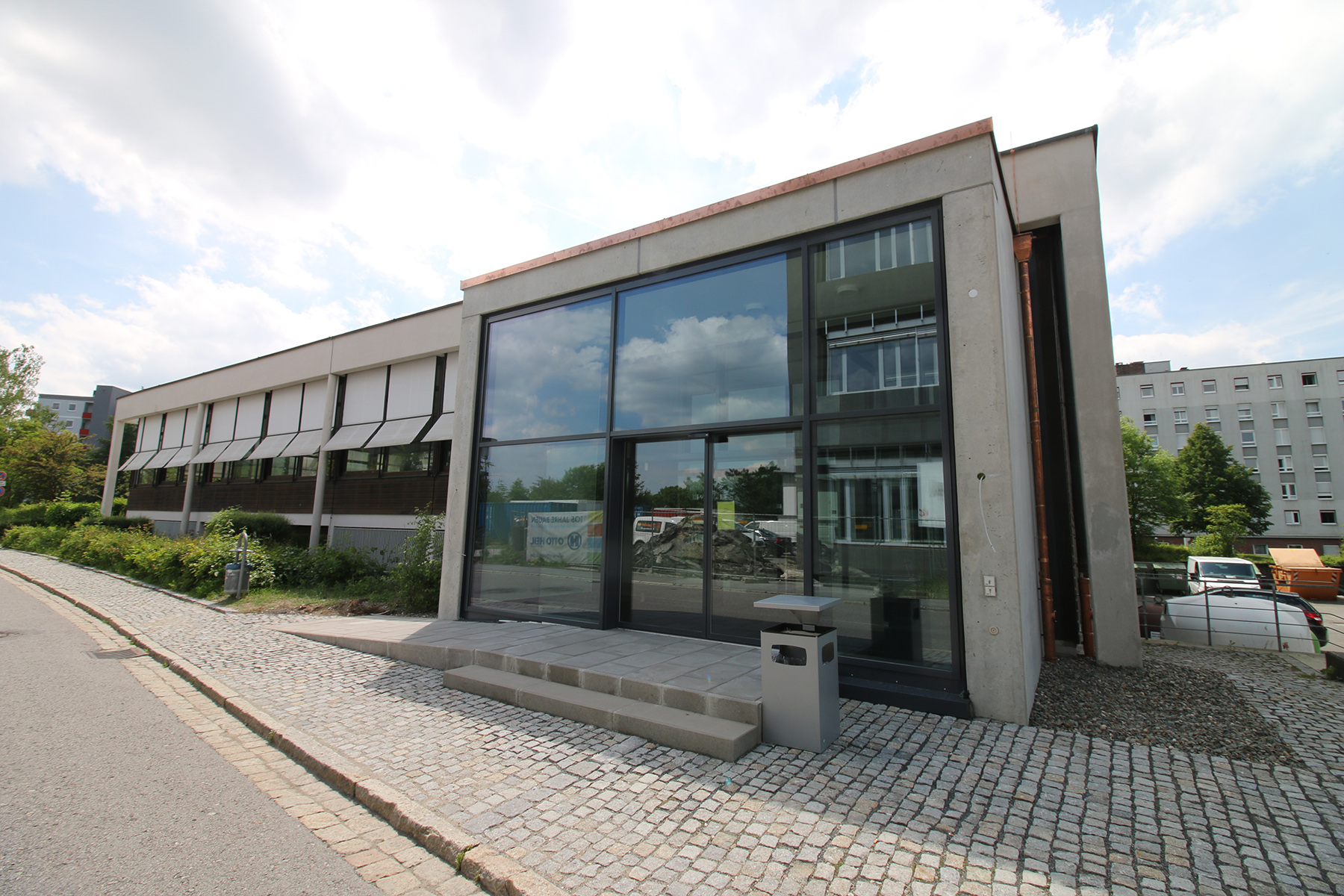 Der Campus der Hochschule Coburg verfügt über ein modernes Gebäude mit großen Glasfenstern, die das Sonnenlicht reflektieren. Ein gepflasterter Weg mit einer kleinen Treppe führt elegant zum Eingang, umgeben von üppigem Grün und eingebettet zwischen benachbarten Gebäuden unter einem teilweise bewölkten Himmel.