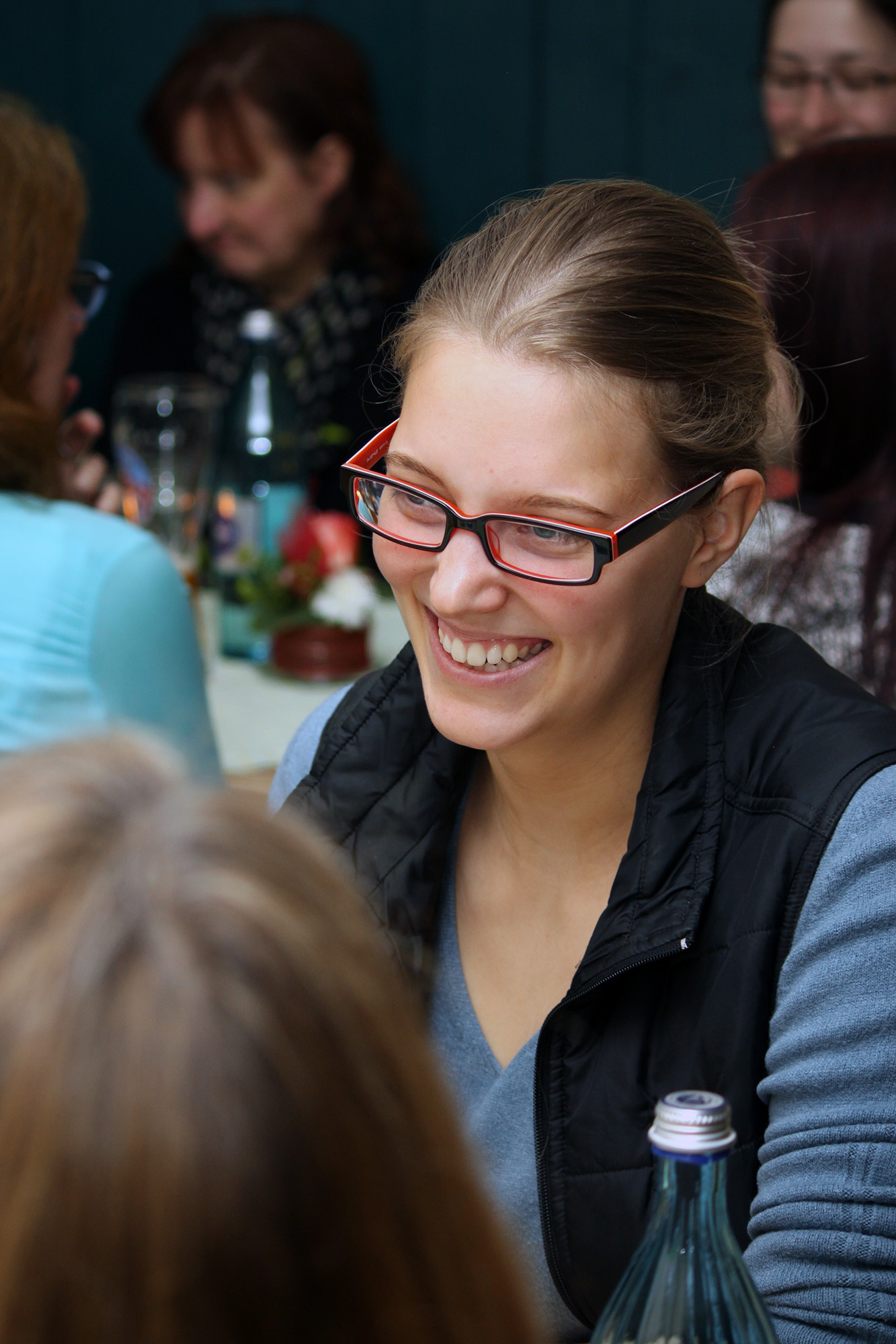 Eine Frau mit Brille und schwarzer Weste sitzt lächelnd an einem Tisch in geselliger Runde, vielleicht nach einer spannenden Vorlesung an der Hochschule Coburg. Im Hintergrund unterhalten sich andere Leute, ihre Diskussionen sind lebhaft. Der Tisch ist mit Flaschen und Gläsern geschmückt, was die warme Atmosphäre widerspiegelt.