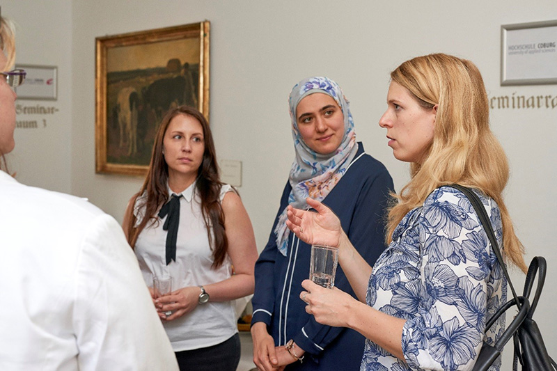 Bei einer Veranstaltung in der Hochschule Coburg unterhalten sich vier Frauen. Eine trägt ein geblümtes Top, eine andere ein Kopftuch und eine dritte eine weiße Bluse mit schwarzer Krawatte. An der Wand hinter ihnen ist ein gerahmtes Gemälde zu sehen. Sie halten Gläser in den Händen, was auf eine lebhafte gesellige Runde schließen lässt.