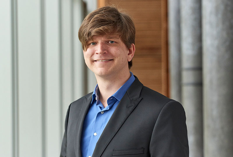 Eine Person mit hellbraunem Haar lächelt, während sie einen schwarzen Blazer und ein blaues Hemd trägt und eine Aura ausstrahlt, die an den lebendigen akademischen Geist der Hochschule Coburg erinnert. Der Hintergrund besteht aus hellgrauen Wänden und einer Holzverkleidung.