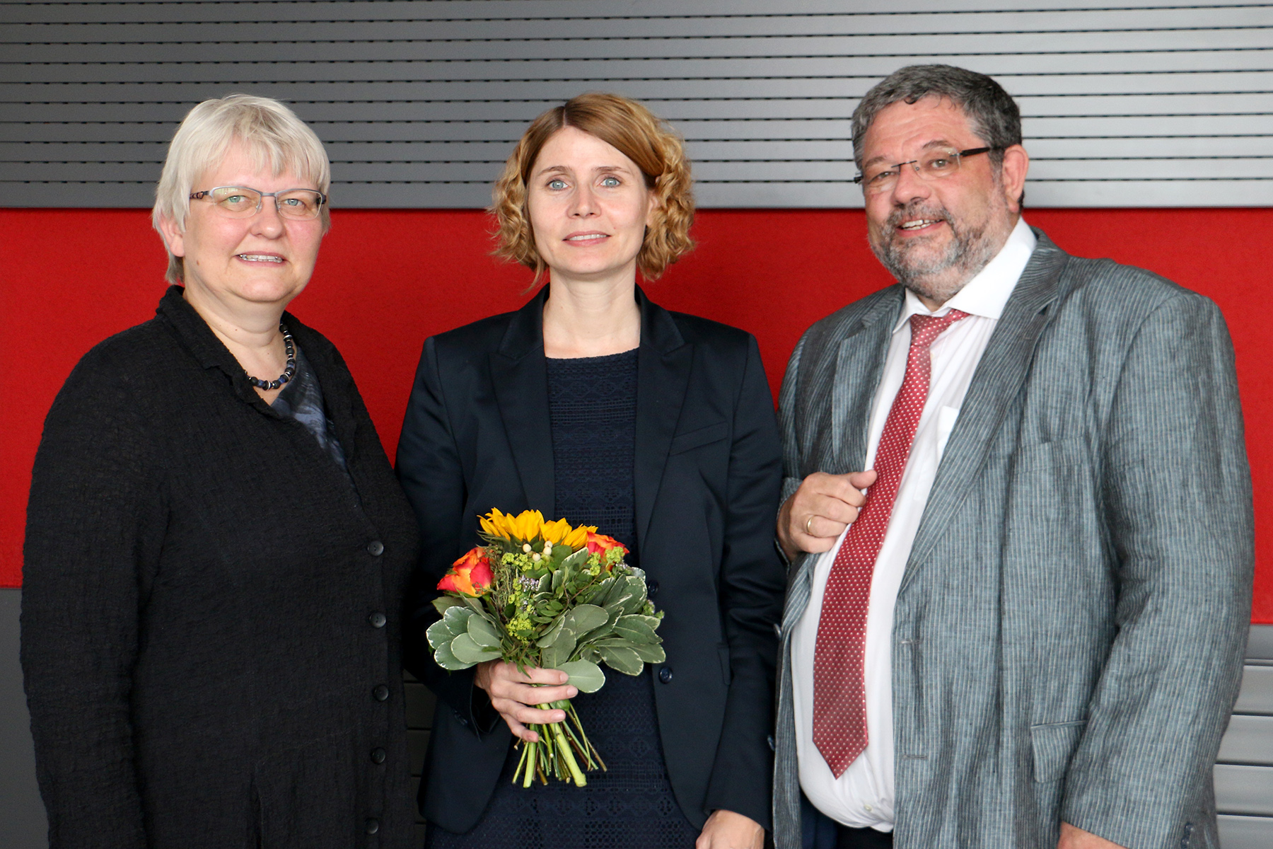 Drei Personen stehen zusammen in einem Innenbereich vor einem rot-grauen Hintergrund. Die Person in der Mitte, Vertreterin der Hochschule Coburg, hält einen Blumenstrauß in der Hand. Sie lächeln alle und sind in formeller Kleidung gekleidet.