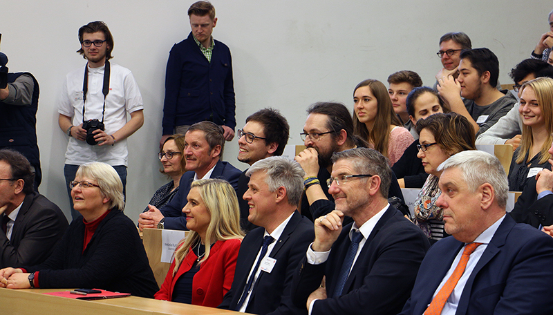 Im Publikum der Hochschule Coburg sitzt eine bunt gemischte Gruppe von Menschen, manche lächeln und sind interessiert, andere wirken aufmerksam. Eine Person im Hintergrund hält eine Kamera und hält das Geschehen fest. Die Kulisse scheint eine Vorlesung oder Präsentation zu sein.