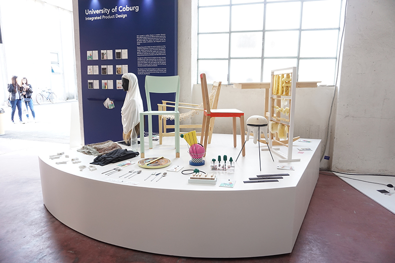 Eine Ausstellung moderner Möbel und Haushaltsgegenstände auf einer weißen Plattform bei einer Ausstellung. Im Hintergrund ist ein Schild der Hochschule Coburg, Integriertes Produktdesign, zu sehen. Im Hintergrund sind Besucher zu sehen.