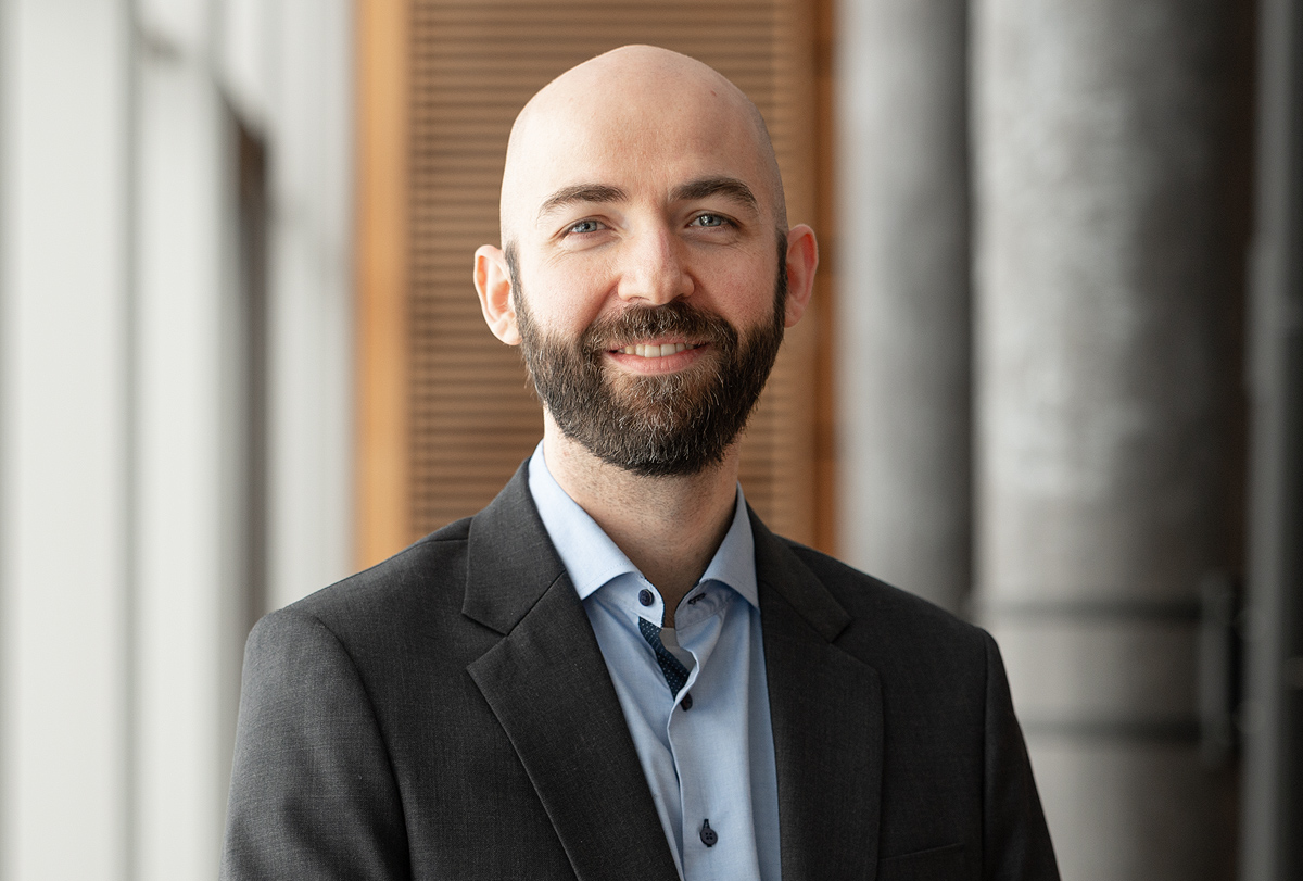 A bald man with a beard is smiling and wearing a dark suit and light blue shirt, standing indoors in front of Hochschule Coburg's signature wooden panels and concrete wall, with natural light streaming in from the left.