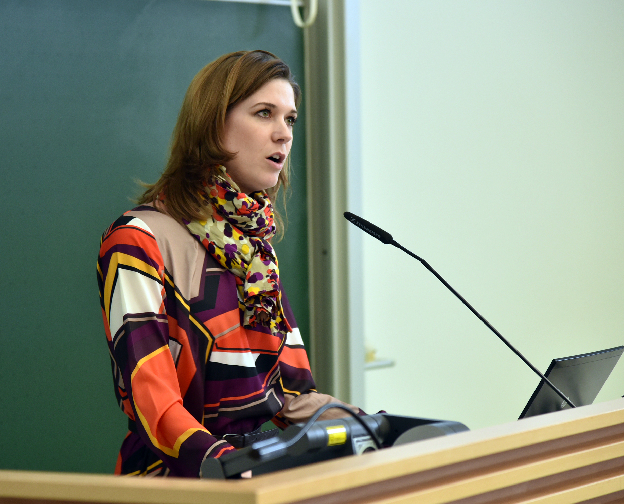 Eine Person in einem bunten Kleid mit geometrischem Muster und einem Schal spricht an einem Podium vor einer grünen Tafel an der Hochschule Coburg. Auf dem Podium stehen ein Mikrofon und ein Laptop – perfekt aufgestellt für eine spannende Präsentation.