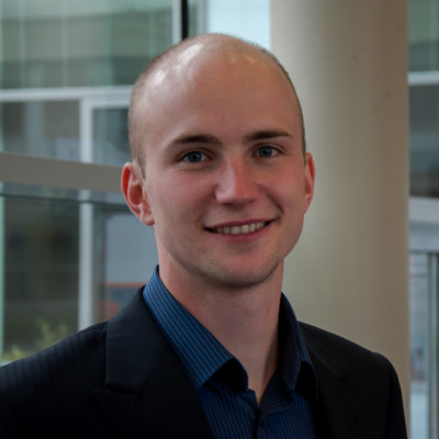 Eine lächelnde Person in einem dunklen Blazer und einem blauen Hemd steht in einem Innenbereich der Hochschule Coburg vor großen Fenstern und einer Säule. Im Hintergrund ist ein modernes Interieur mit verschwommenen architektonischen Details zu sehen.