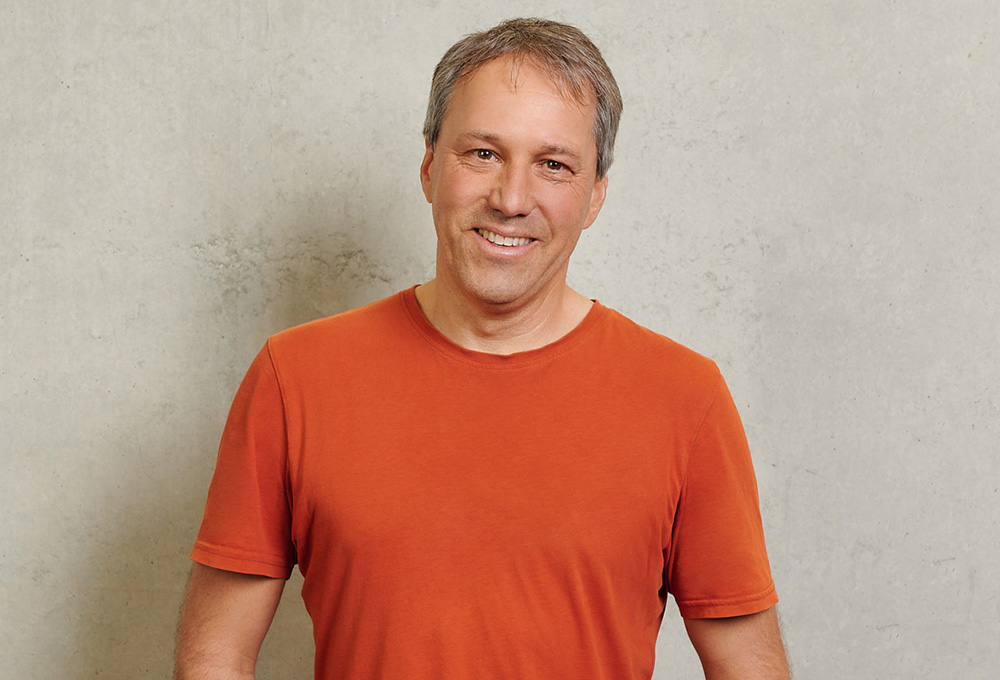 Ein Mann trägt ein orangefarbenes T-Shirt, auf dem stolz das Logo der Hochschule Coburg zu sehen ist, lächelt, während er vor einer schlichten hellen Wand steht.