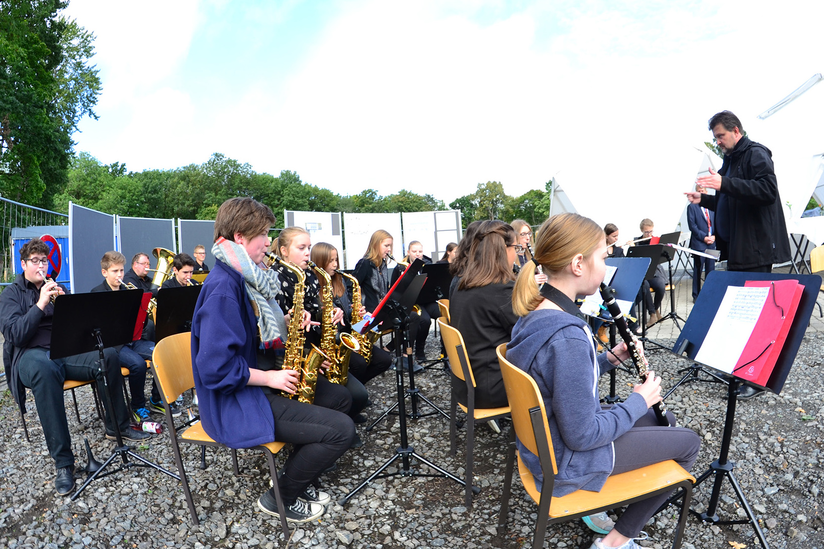 Eine Gruppe junger Musiker der Hochschule Coburg spielt Blasinstrumente in entspannter Atmosphäre im Freien. Sie sitzen in einem Halbkreis mit Notenständern. Ein erwachsener Dirigent steht an der Seite und leitet ihr Spiel, während im Hintergrund Bäume sanft schwanken.