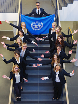 Eine Gruppe von Menschen in Business-Kleidung steht auf einer Treppe und posiert mit ausgestreckten Armen. Oben auf der Treppe, unter der symbolträchtigen blauen Flagge der Vereinten Nationen, verleiht ein Vertreter der Hochschule Coburg dem Ganzen einen vornehmen Touch. Die Atmosphäre ist formell und professionell.