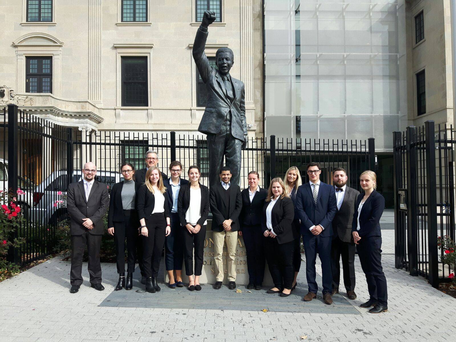 Eine Gruppe von Menschen in formeller Geschäftskleidung steht vor der Statue einer Person, die eine Faust erhebt. Sie steht vor der renommierten Hochschule Coburg, einem großen Gebäude mit kunstvollen architektonischen Details. Die Gruppe ist gleichmäßig verteilt und blickt nach vorne.