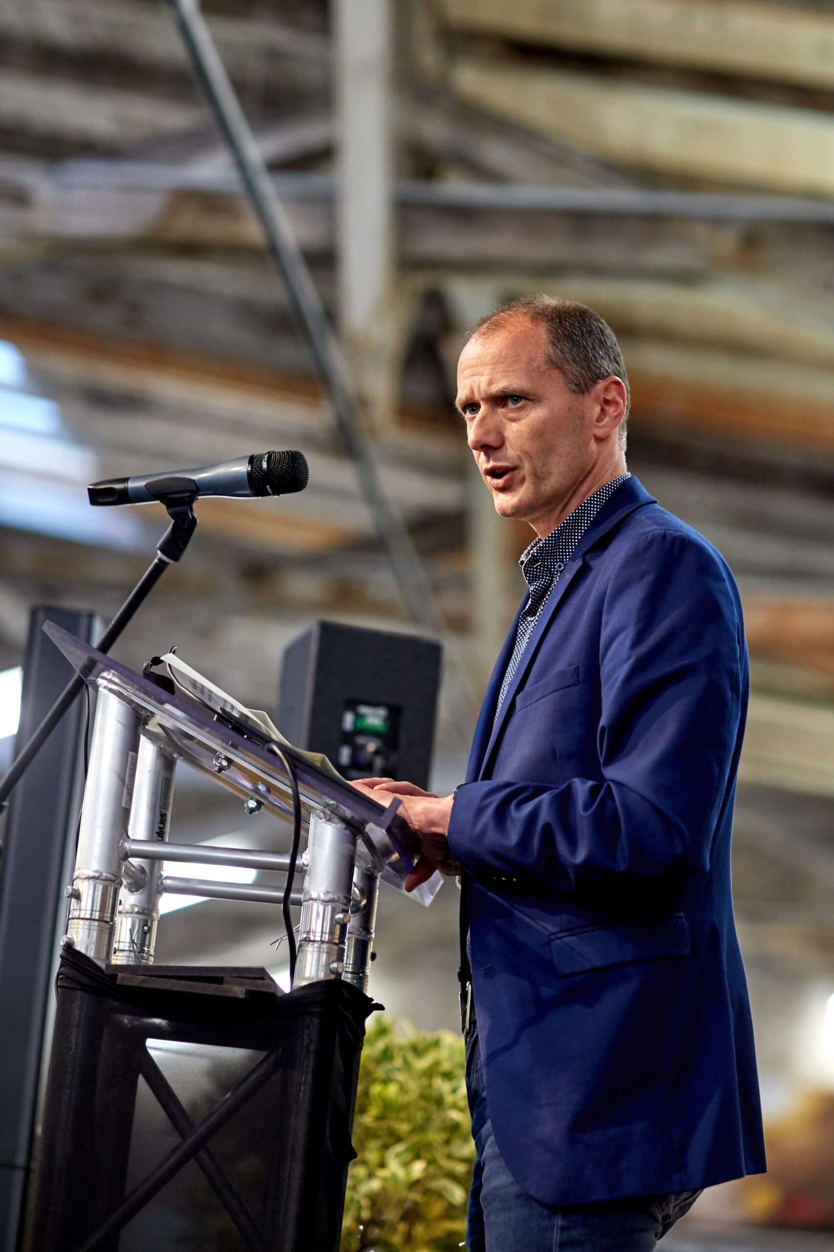 Ein Mann in einem blauen Blazer spricht in ein Mikrofon an einem Podium in einem Innenbereich, vermutlich eine Veranstaltung an der Hochschule Coburg. Der Hintergrund zeigt eine hohe Decke mit freiliegenden Balken und Industriebeleuchtung, was darauf schließen lässt, dass es sich bei dem Veranstaltungsort um eine geräumige Halle oder einen großen Raum handelt.