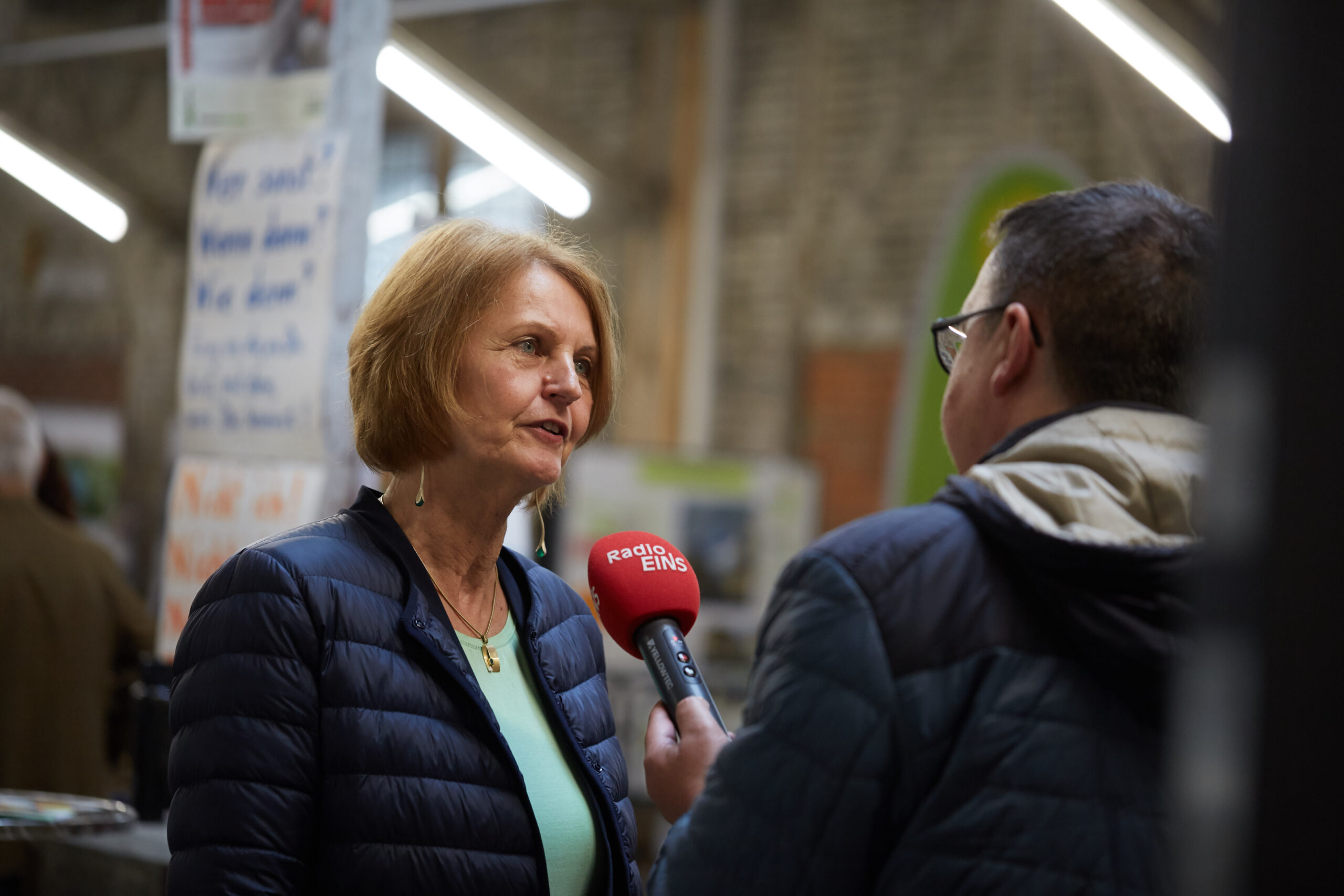 Eine Frau in dunkler Jacke wird von einem Mann mit Brille und Kapuzenjacke in einem Gebäude interviewt, das wie die Hochschule Coburg aussieht. Der Mann hält ein rotes Mikrofon mit der Aufschrift „Radio Eins“ unter dem hellen Deckenlicht, im Hintergrund sind Plakate zu sehen.