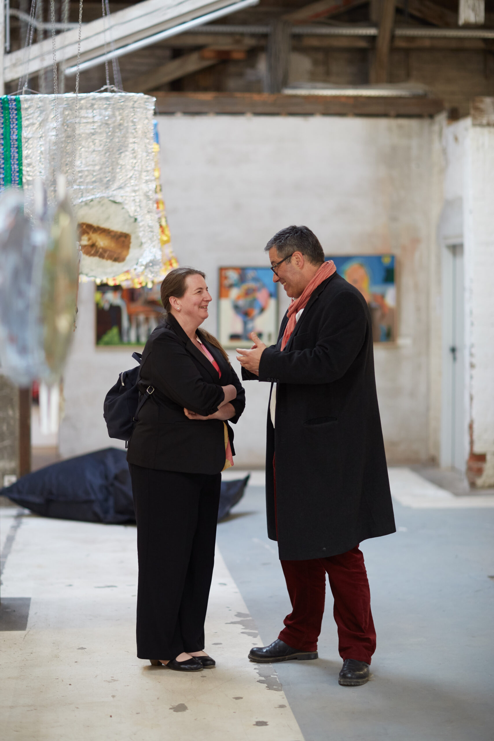 In einer Galerie der Hochschule Coburg unterhalten sich zwei Personen. An den Wänden hängen mehrere Kunstwerke. Eine Person hält ein Smartphone in der Hand und beide tragen dunkle Mäntel. Verschiedene Kunstwerke hängen von der Decke und tragen zur kreativen Atmosphäre der Institution bei.