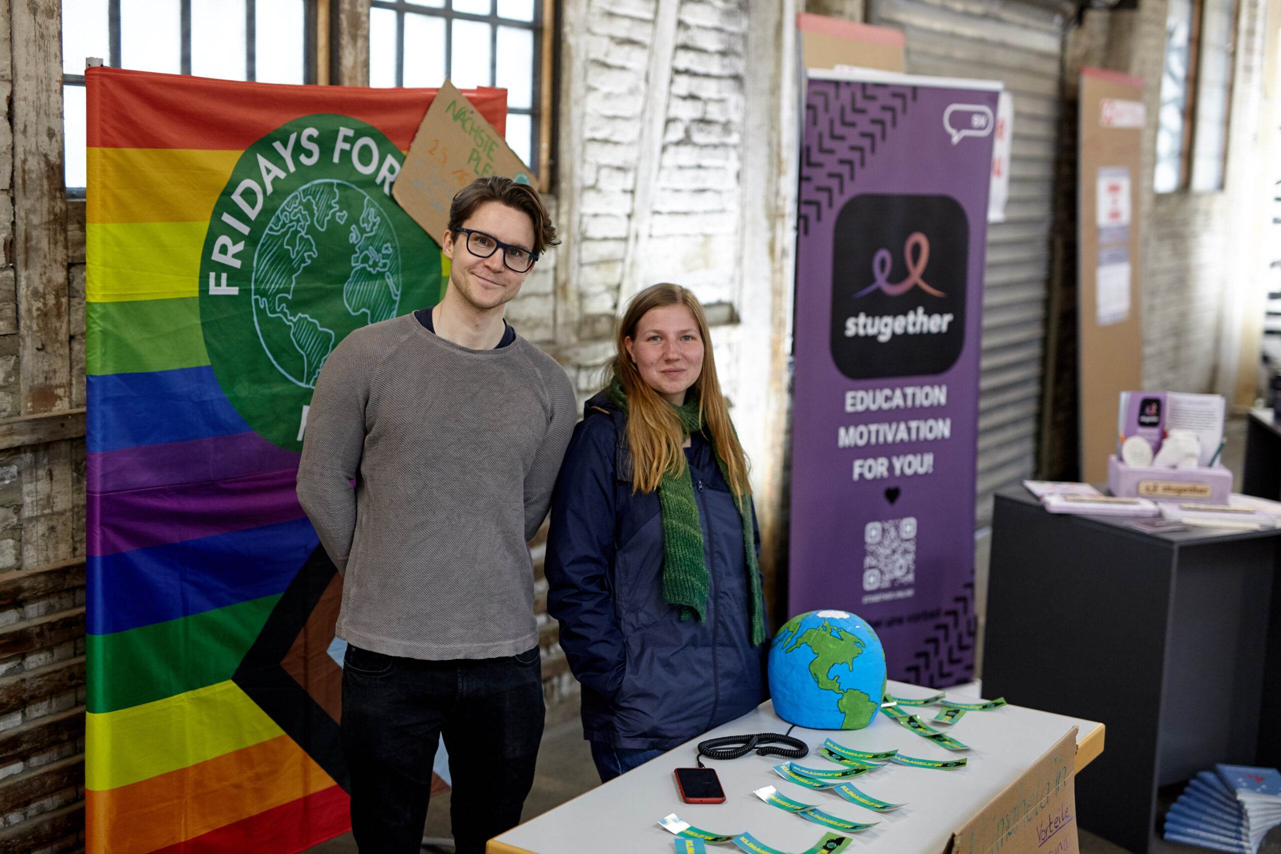 Zwei Personen stehen an einem Stand mit einem Regenbogenbanner mit der Aufschrift „Fridays for Future“ und einem kleinen Globus und werben für Umweltaktivismus neben den Initiativen der Hochschule Coburg. Ein „Stugether“-Schild in der Nähe wirbt für Bildung und Motivation, während auf den Tischen Broschüren und andere interessante Materialien ausgestellt sind.