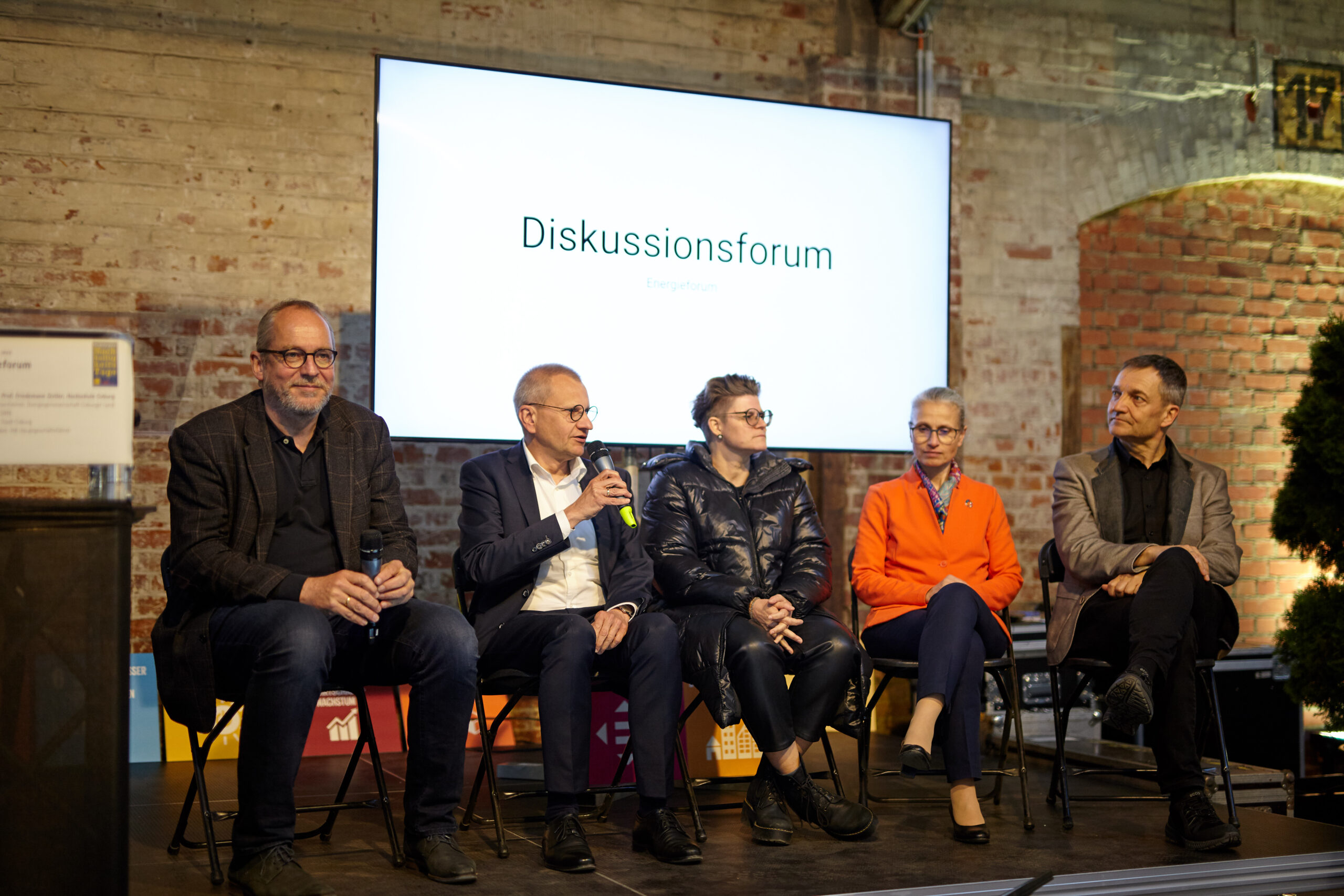 Fünf Personen sitzen auf der Bühne eines Diskussionsforums an der Hochschule Coburg. Sie sitzen vor einer großen Leinwand mit der Aufschrift „Diskussionsforum“. Die Gruppe besteht aus drei Männern und zwei Frauen, alle in Business-Casual-Kleidung gekleidet und bereit für den akademischen Dialog.