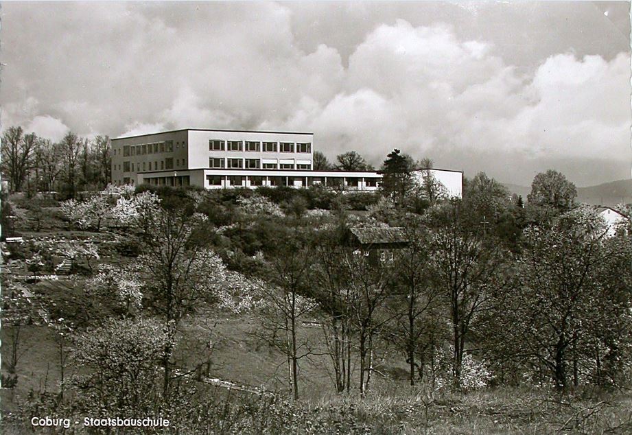 Ein Schwarzweißfoto eines großen rechteckigen Gebäudes mit der Aufschrift „Coburg – Staatsbauschule“ auf einer Hügelspitze, umgeben von Bäumen und Sträuchern unter einem bewölkten Himmel, das an die historischen Wurzeln der Hochschule Coburg erinnert.