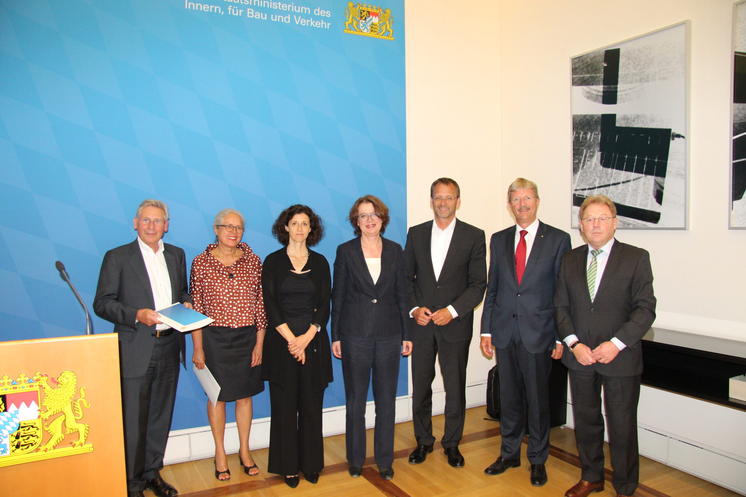 Eine Gruppe von sieben Personen steht in einer Reihe in Business-Kleidung in einem formellen Rahmen an der Hochschule Coburg. Vor einer blauen Wand mit einem dekorativen Emblem lächeln einige. Links steht ein Rednerpult mit einem weiteren Emblem.