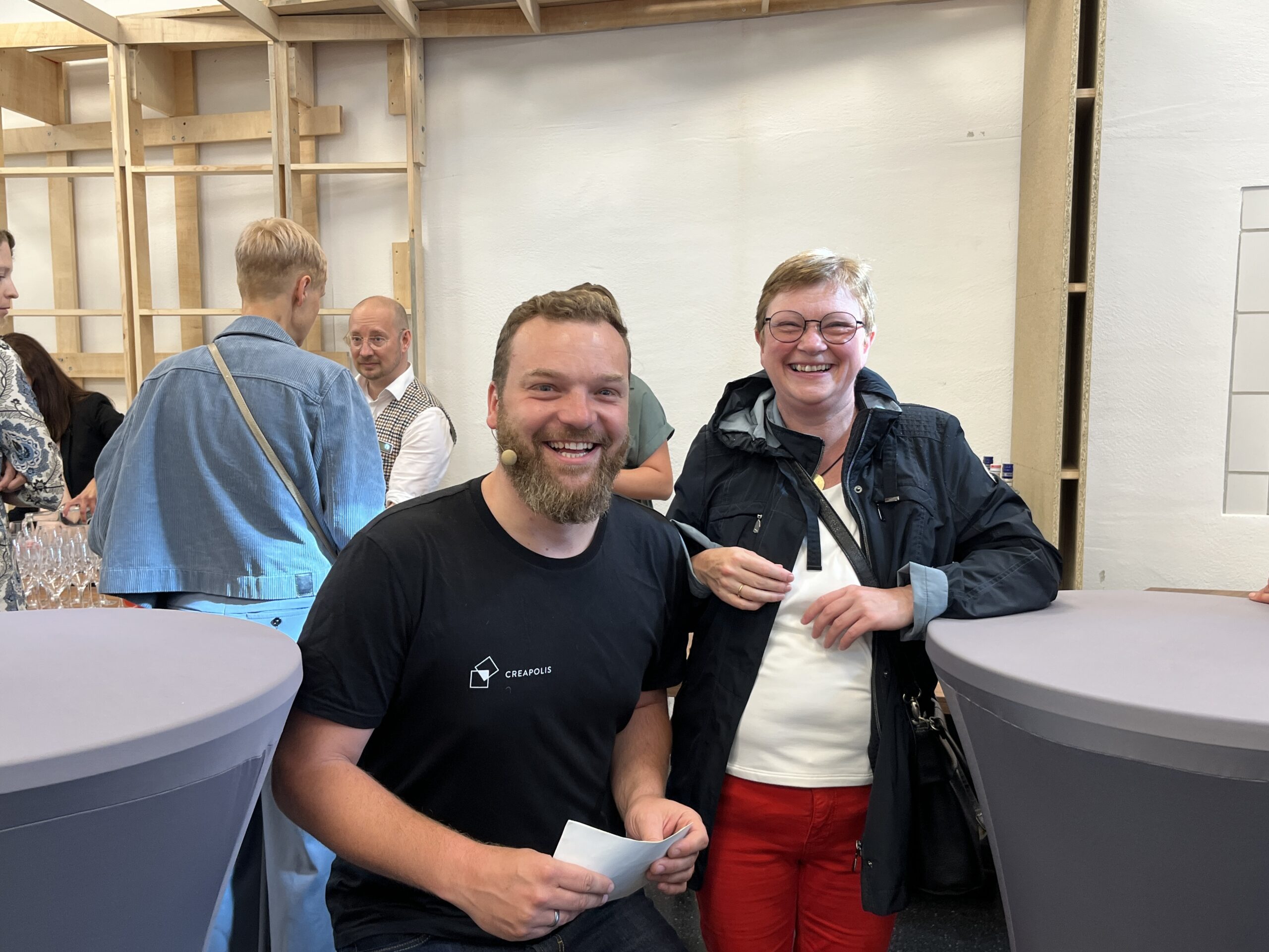 Ein Mann sitzt und lächelt neben einer Frau, die steht. Beide strahlen bei einer Veranstaltung der Hochschule Coburg eine fröhliche Stimmung aus. Um sie herum stehen Stehtische, im Hintergrund mischen sich andere Leute zwanglos unter die Leute.