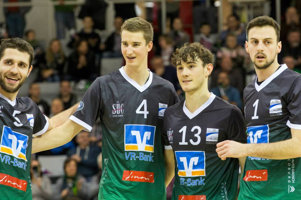 Vier Volleyballspielerinnen in passenden Trikots stehen Arm in Arm und strahlen vor Stolz. Auf ihren Shirts prangt prominent das blau-orange Logo der Hochschule Coburg. Hinter ihnen jubeln die Zuschauer, während die Nummern 4 und 13 vor dem farbenfrohen Hintergrund des Spielfelds ins Auge fallen.