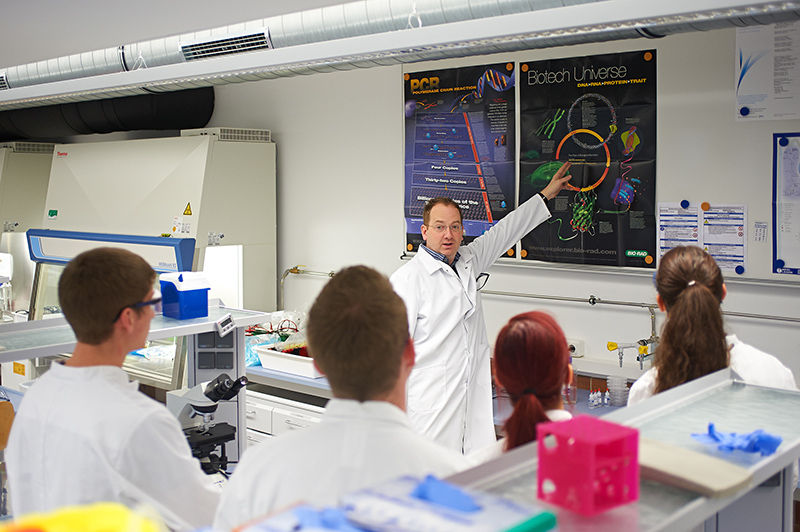 Ein Wissenschaftler der Hochschule Coburg im Laborkittel hält vor einer Gruppe von fünf Personen in einem Labor einen Vortrag. Er zeigt auf ein buntes Poster mit dem Titel „Biotech Universe“. Die Anwesenden, ebenfalls in Laborkitteln, sind von verschiedenen Laborgeräten umgeben.