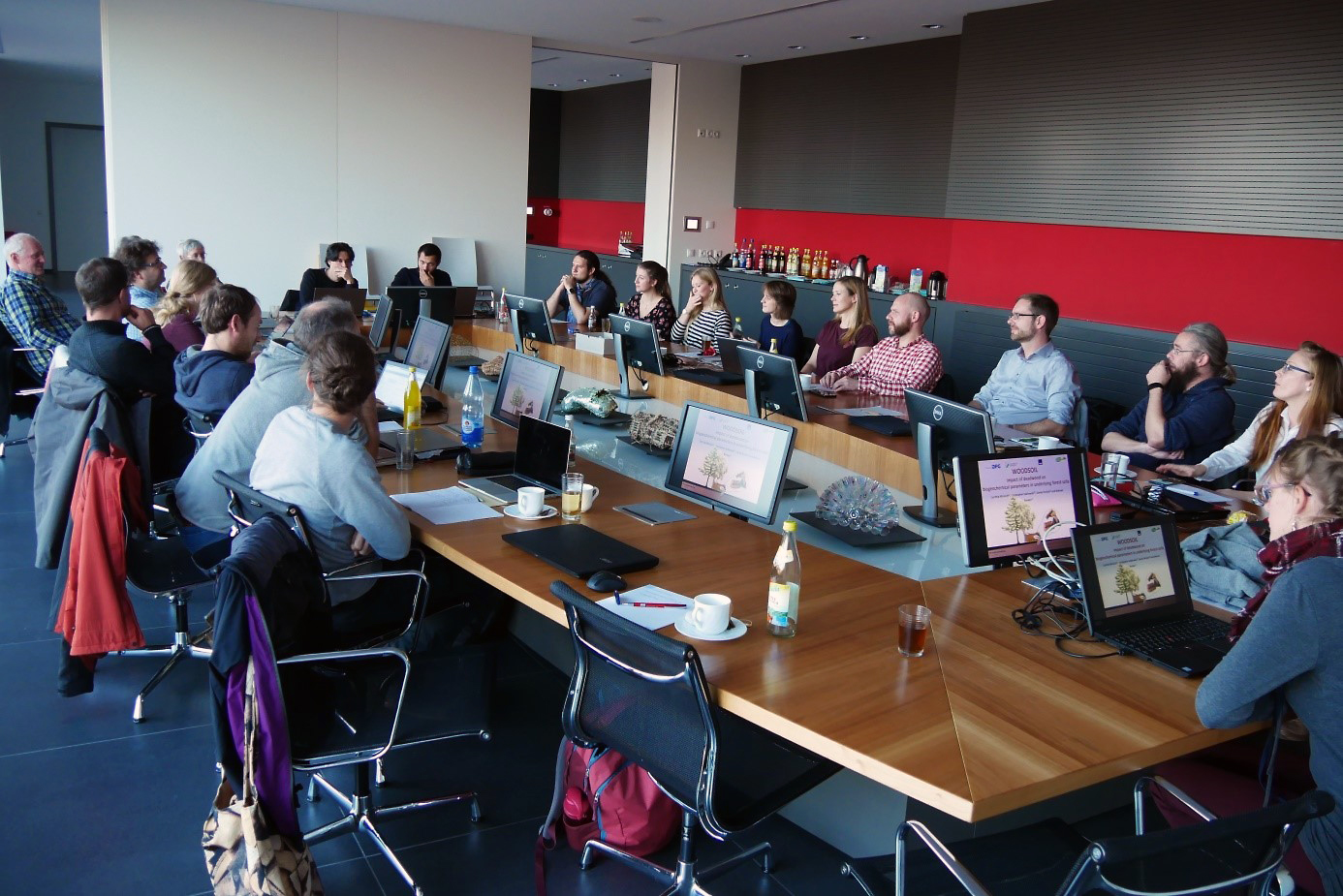 Eine Gruppe von Mitarbeitern der Hochschule Coburg sitzt in einem modernen Büro um einen großen U-förmigen Konferenztisch. Jeder hat einen Laptop, auf Bildschirmen laufen Präsentationsfolien. Der Raum ist in Rot und Grau gehalten, im hinteren Bereich gibt es Erfrischungen.