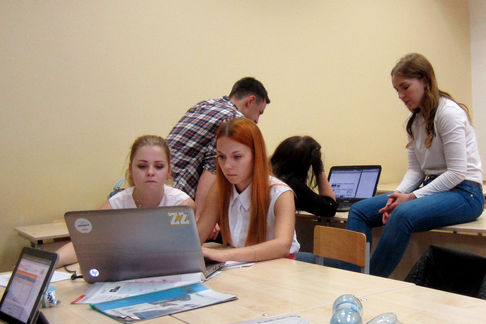 In einem Hörsaal der Hochschule Coburg arbeitet eine Gruppe junger Erwachsener fleißig zusammen. Zwei Frauen sitzen an einem Tisch konzentriert auf einen Laptop, während ein anderer Mann und zwei Frauen in der Nähe mit Laptops und Papieren herumhantieren. So entsteht eine Atmosphäre der Teamarbeit und des Lernens.