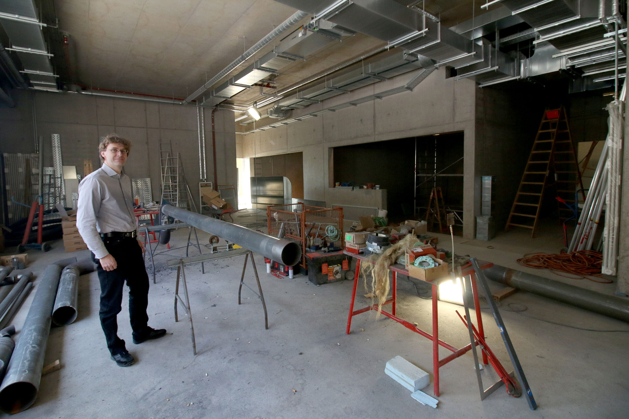 Ein Mann steht in einem teilweise fertiggestellten Industriegebäude der Hochschule Coburg mit freiliegenden Leitungen und Betonwänden. Verschiedene Baumaterialien und Werkzeuge liegen verstreut herum, und in einer Ecke ist eine Leiter zu sehen.