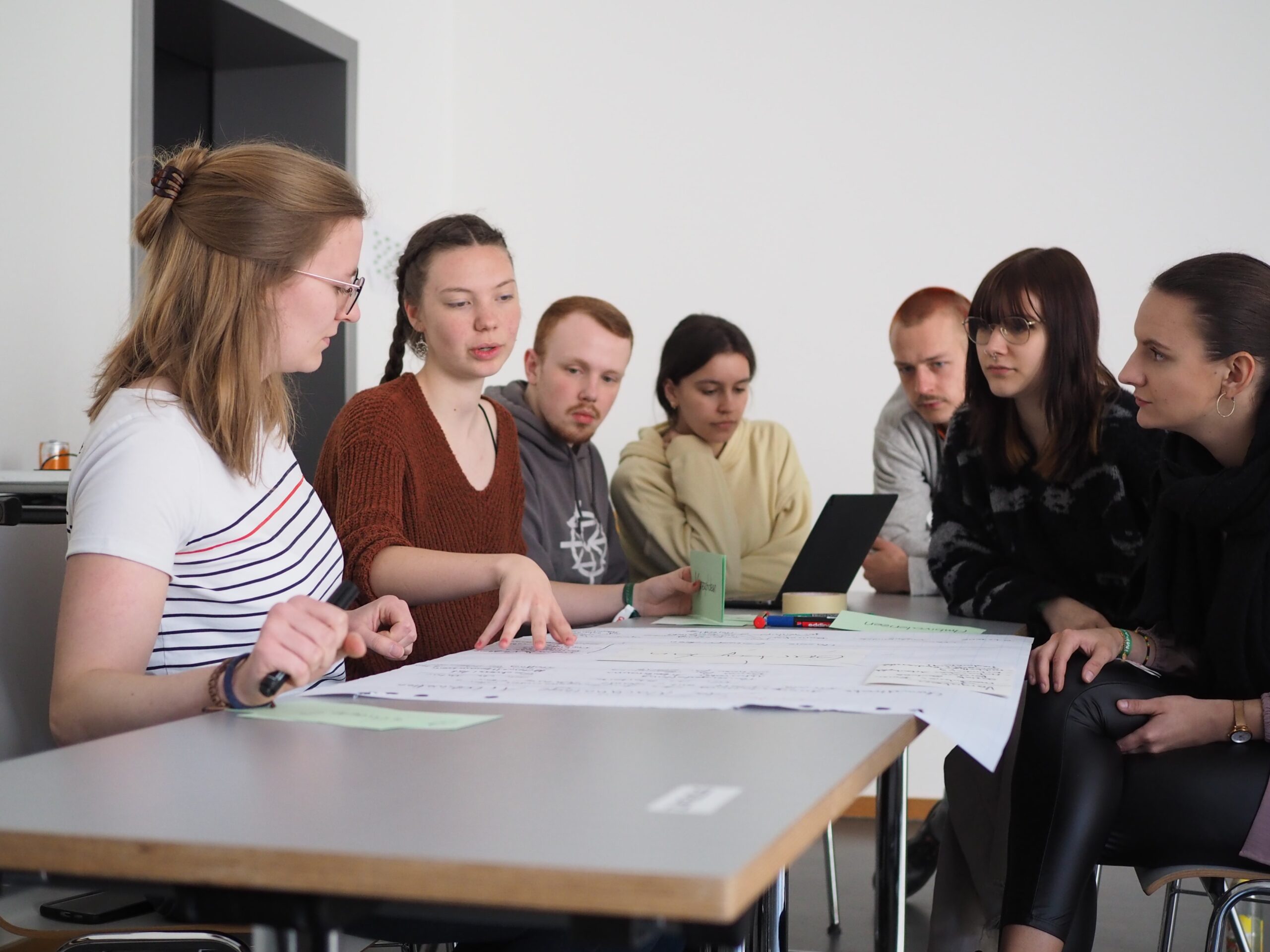 Eine Gruppe von sieben Personen sitzt an einem Tisch in der Hochschule Coburg und diskutiert. Einige studieren große Blätter mit Notizen, einer sitzt am Laptop. Der Raum ist hell und minimalistisch.