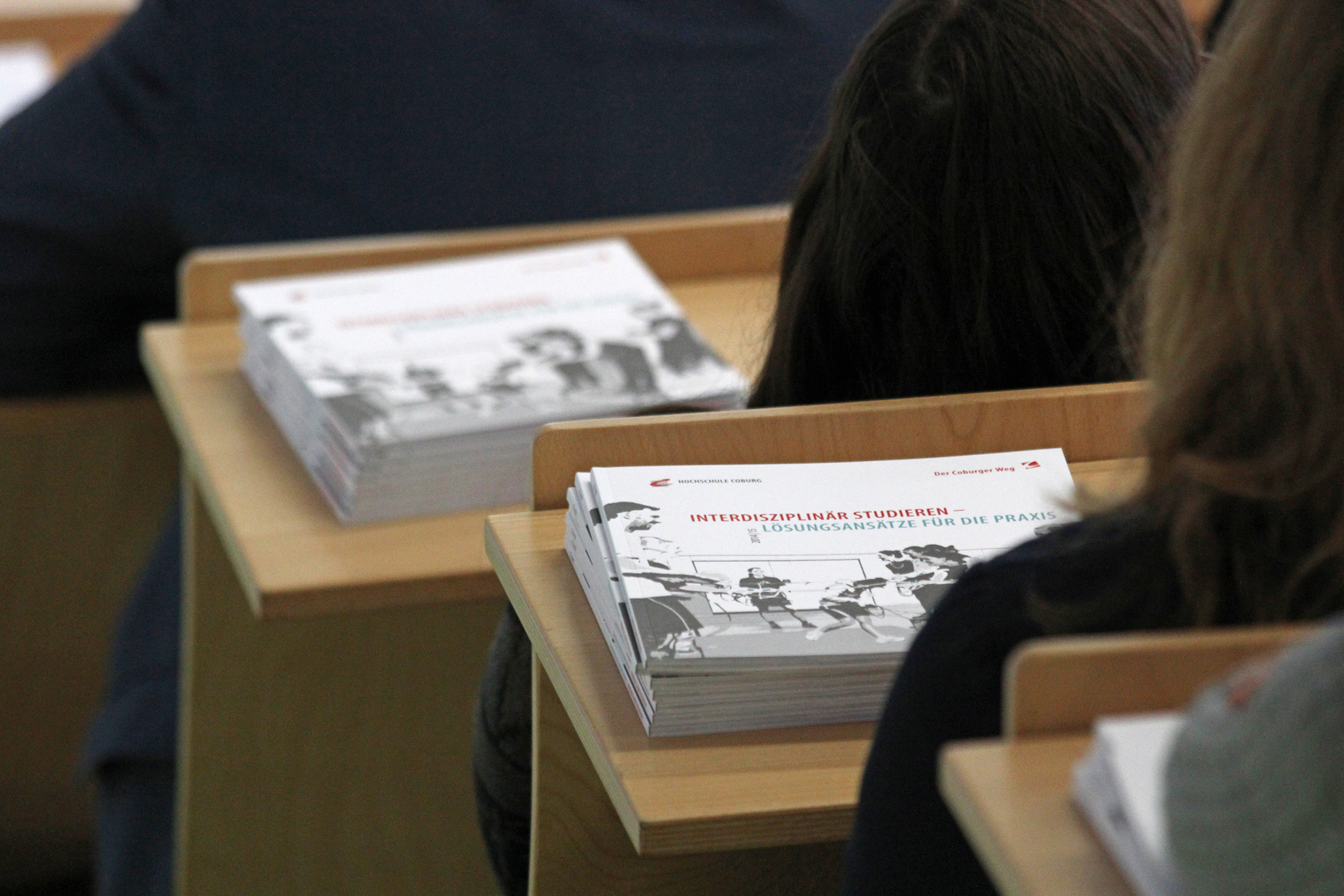 Stapelweise Broschüren mit dem Titel „Interdisziplinär Studieren“ liegen auf Holztischen in einem Hörsaal der Hochschule Coburg. Einige Zuhörer sitzen und blicken nach vorne.