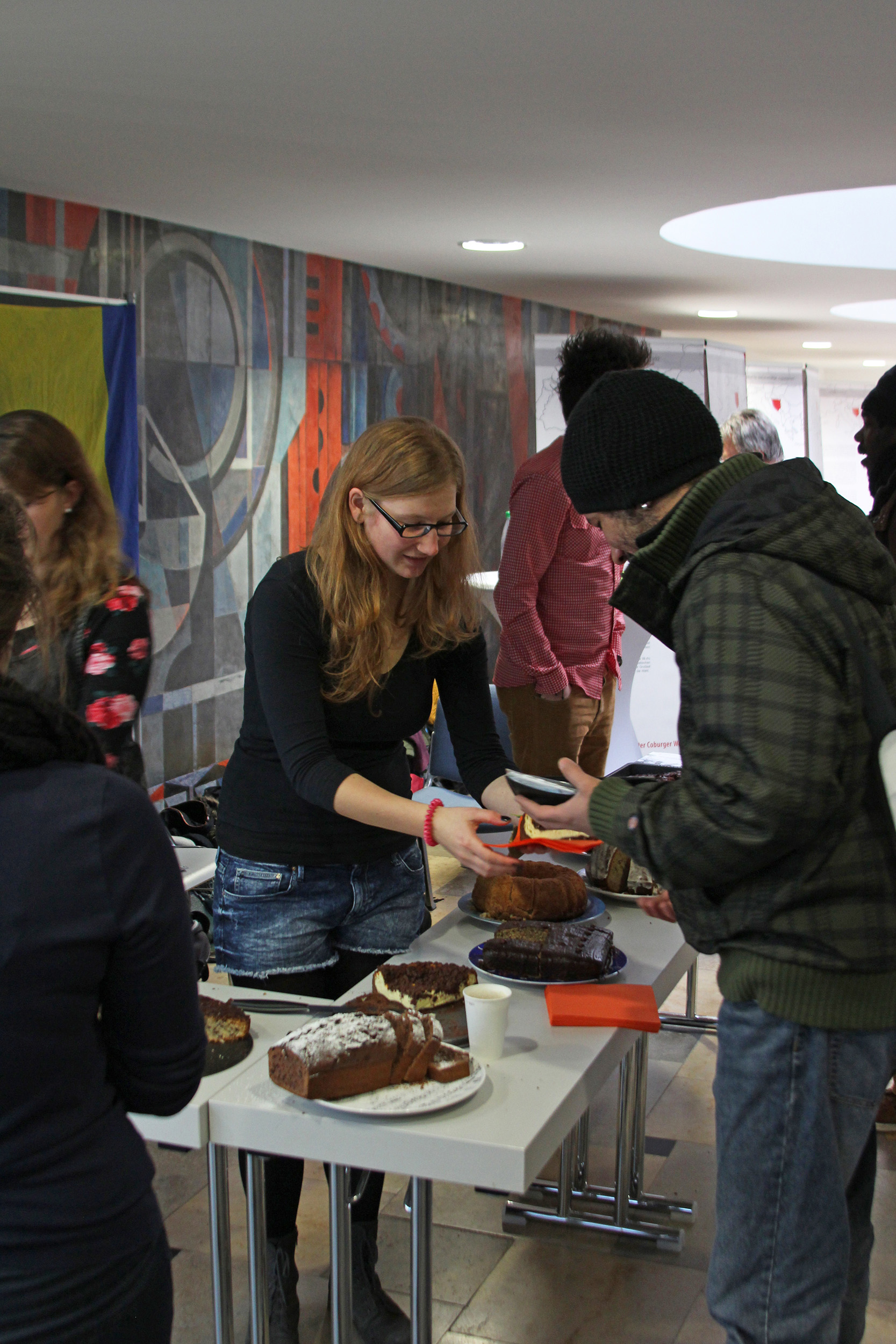 Bei einer Veranstaltung der Hochschule Coburg serviert eine Frau mit Brille einer Person mit Beanie an einem Kuchenverkaufstisch im Innenbereich Kuchen. Verschiedene Kuchen werden ausgestellt, während sich im Hintergrund bei einem farbenfrohen Wandgemälde andere Teilnehmer versammeln.