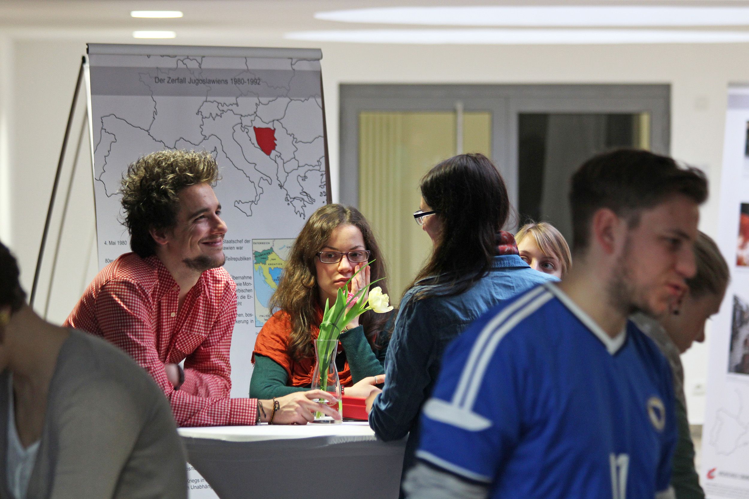 Bei einer Veranstaltung der Hochschule Coburg unterhält sich eine Gruppe von Menschen angeregt an einem Tisch. Ein Mann und eine Frau am Tisch bewundern eine Vase mit Blumen. Im Hintergrund ist auf einer Tafel eine Karte zu sehen, die bei den Teilnehmern Neugier und Inspiration weckt.