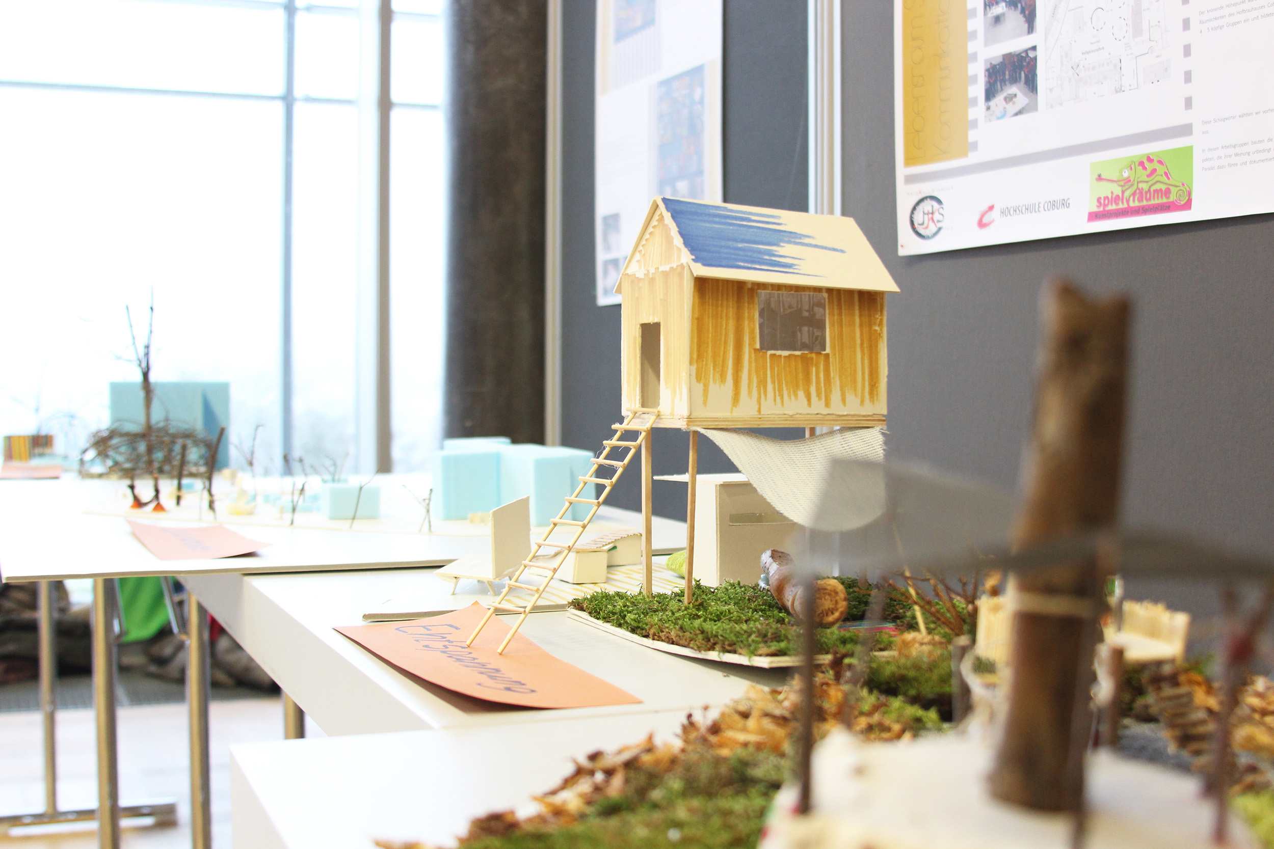 Eine Ausstellung von Miniatur-Architekturmodellen auf weißen Tischen zieht die Besucher der Hochschule Coburg in ihren Bann. Unter ihnen sticht ein kleines Holzhaus auf Stelzen mit einer Leiter hervor. Im Hintergrund sind verschiedene andere Modelle und Informationsplakate zu sehen, die eine Ausstellung oder einen pädagogischen Rahmen suggerieren.