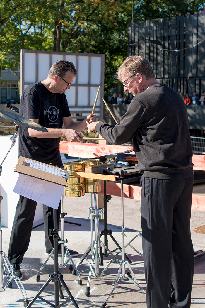 Zwei Männer in schwarzen Hemden spielen im Freien an der Hochschule Coburg Schlaginstrumente. Einer hält Trommelstöcke, während der andere ein Instrument einstellt. Vor ihnen steht ein Notenständer mit Notenblättern. Bäume, eine Menschenmenge und Holzkonstruktionen tragen zur lebhaften Hintergrundszene bei.