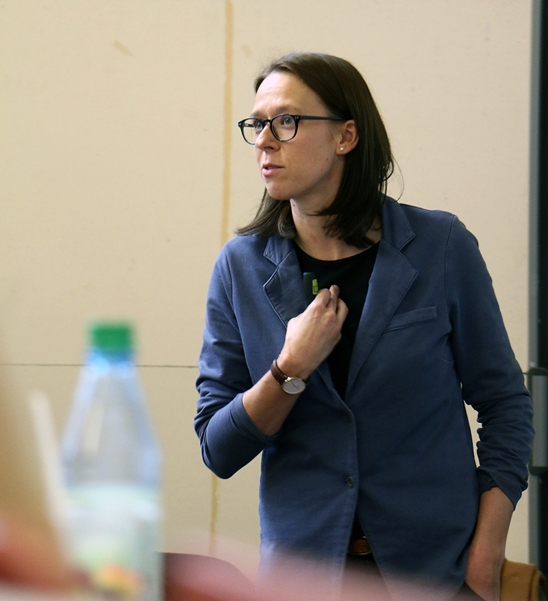 Eine Person mit langen Haaren und Brille steht drinnen und trägt einen blauen Blazer. Sie hält eine Präsentationsfernbedienung in der Hand und scheint einen Vortrag an der Hochschule Coburg zu halten. Im Vordergrund ist eine verschwommene Wasserflasche zu sehen.