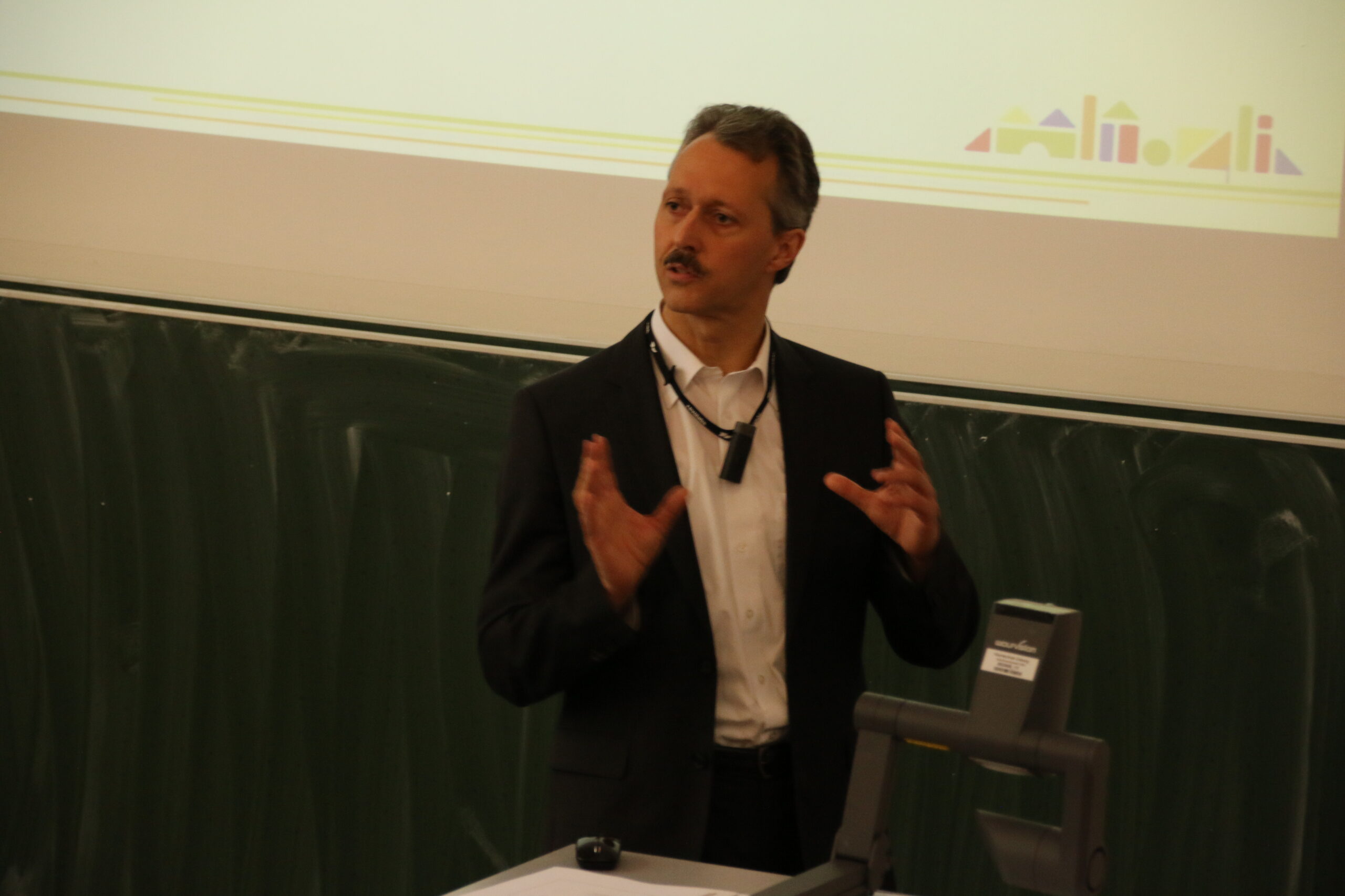 Ein Mann im Anzug gestikuliert, während er an einem Podium in einem Hörsaal spricht. Hinter ihm ist auf einer großen Leinwand neben einer leeren Projektion das Logo der Hochschule Coburg teilweise zu sehen.