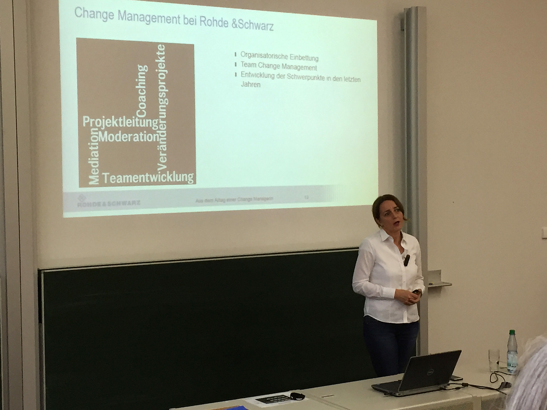 Eine Frau steht vor einem Hörsaal der Hochschule Coburg und hält für Rohde & Schwarz einen Vortrag zum Thema „Change Management“. Auf einer großen Leinwand werden zentrale Themen wie Teamentwicklung und Moderation präsentiert. Im Vordergrund komplettieren eine Tafel und ein Laptop die dynamische Lernumgebung.