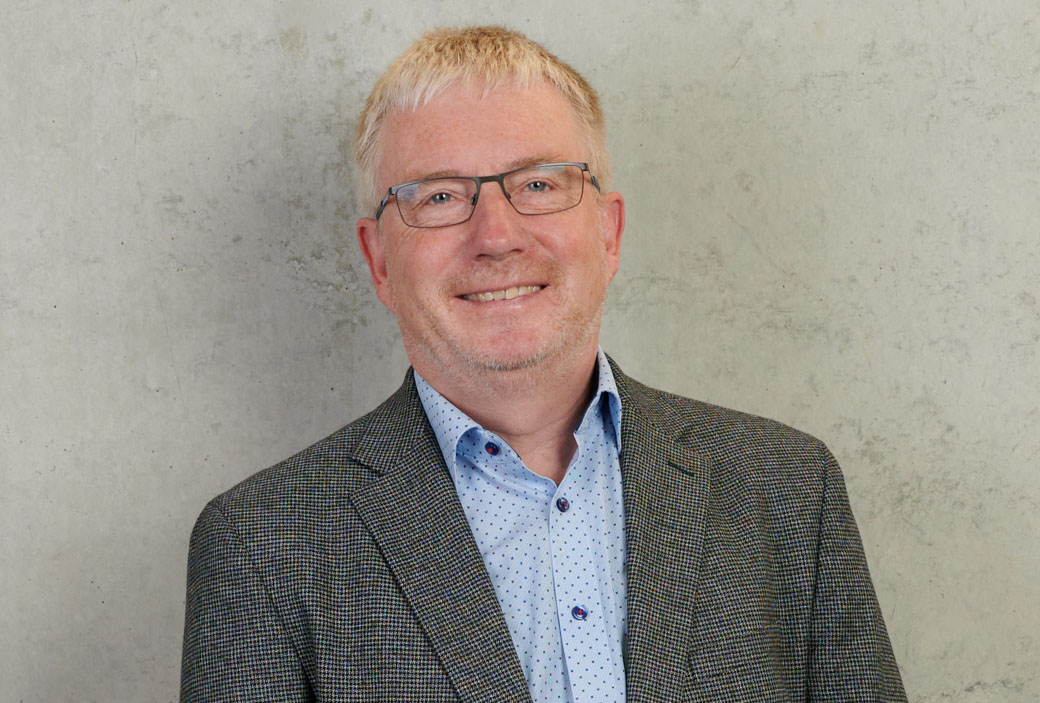 A smiling man with glasses, short light hair, and a beard wears a grey checkered blazer over a light blue patterned shirt. Standing before a plain textured wall, he exudes the confidence of someone affiliated with Hochschule Coburg.