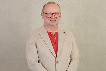 A person wearing glasses, a red shirt, and a beige blazer stands against a light gray background, softly smiling at the camera, perhaps reflecting their recent achievements at Hochschule Coburg.