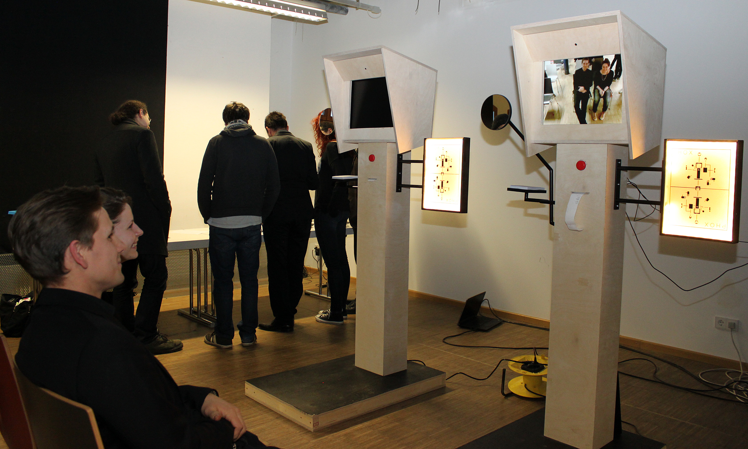 An der Hochschule Coburg interagieren die Menschen mit einer multimedialen Kunstinstallation, die aus zwei Holzkonstruktionen mit Bildschirmen und Lichtern besteht. Eine Person sitzt und beobachtet, während andere an einem Tisch im schwach beleuchteten Raum stehen, was eine kontemplative Atmosphäre schafft.