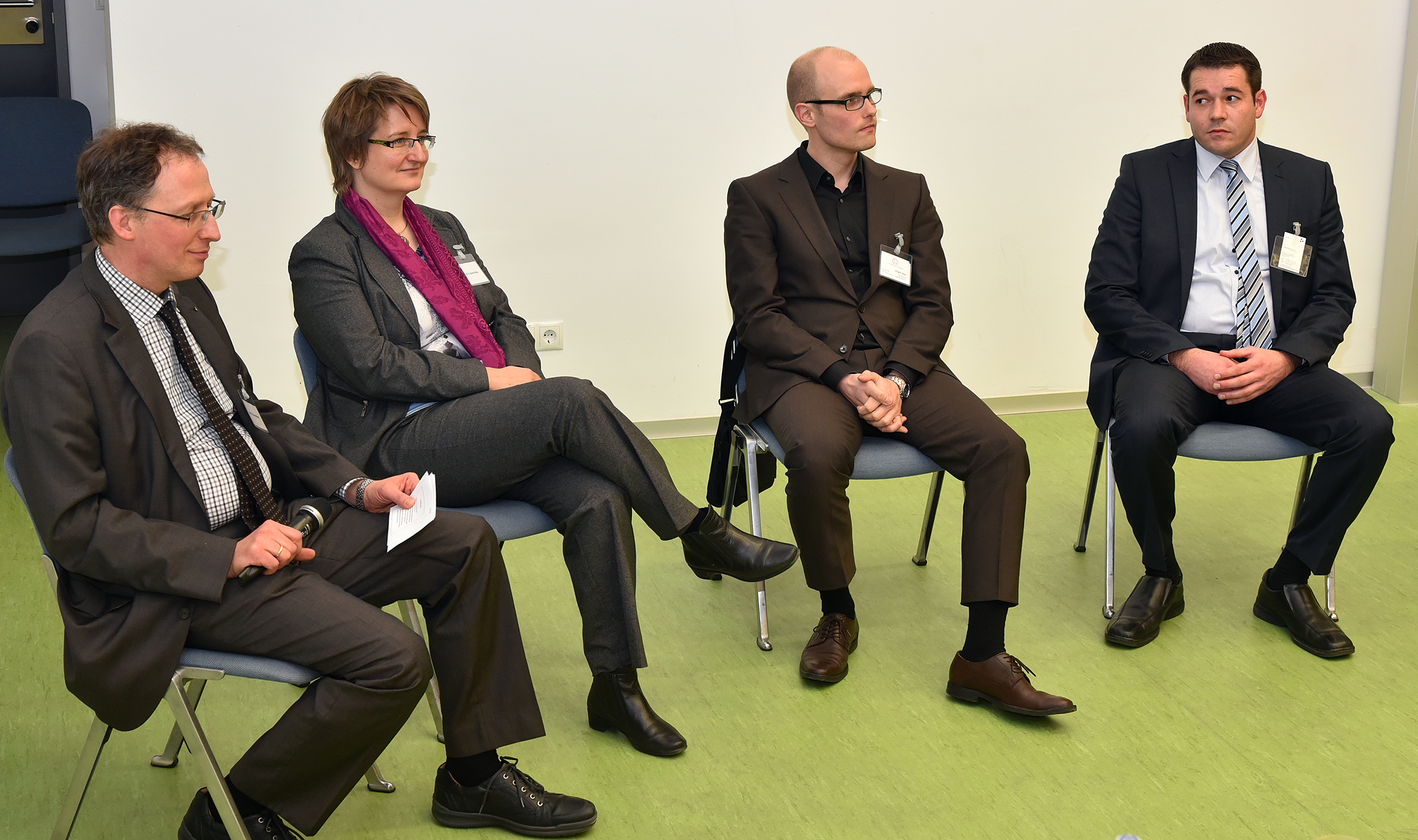 Vier Personen in formeller Kleidung sitzen im Halbkreis auf blauen Stühlen und beteiligen sich aktiv an einer Diskussion. Die Umgebung ähnelt einem Konferenzraum der Hochschule Coburg, wobei eine schlichte weiße Wand als Hintergrund dient.