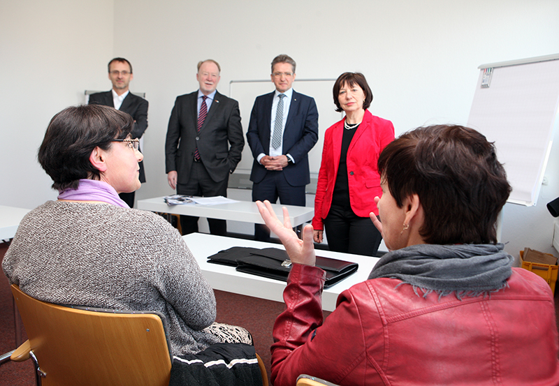 Eine Gruppe von fünf Personen in Business-Kleidung befindet sich in einem Besprechungsraum der Hochschule Coburg. Drei Personen stehen vorne, während zwei mit dem Rücken zur Kamera sitzen. Die Kulisse umfasst Stühle, Schreibtische und ein Whiteboard. Eine Person gestikuliert beim Sprechen.