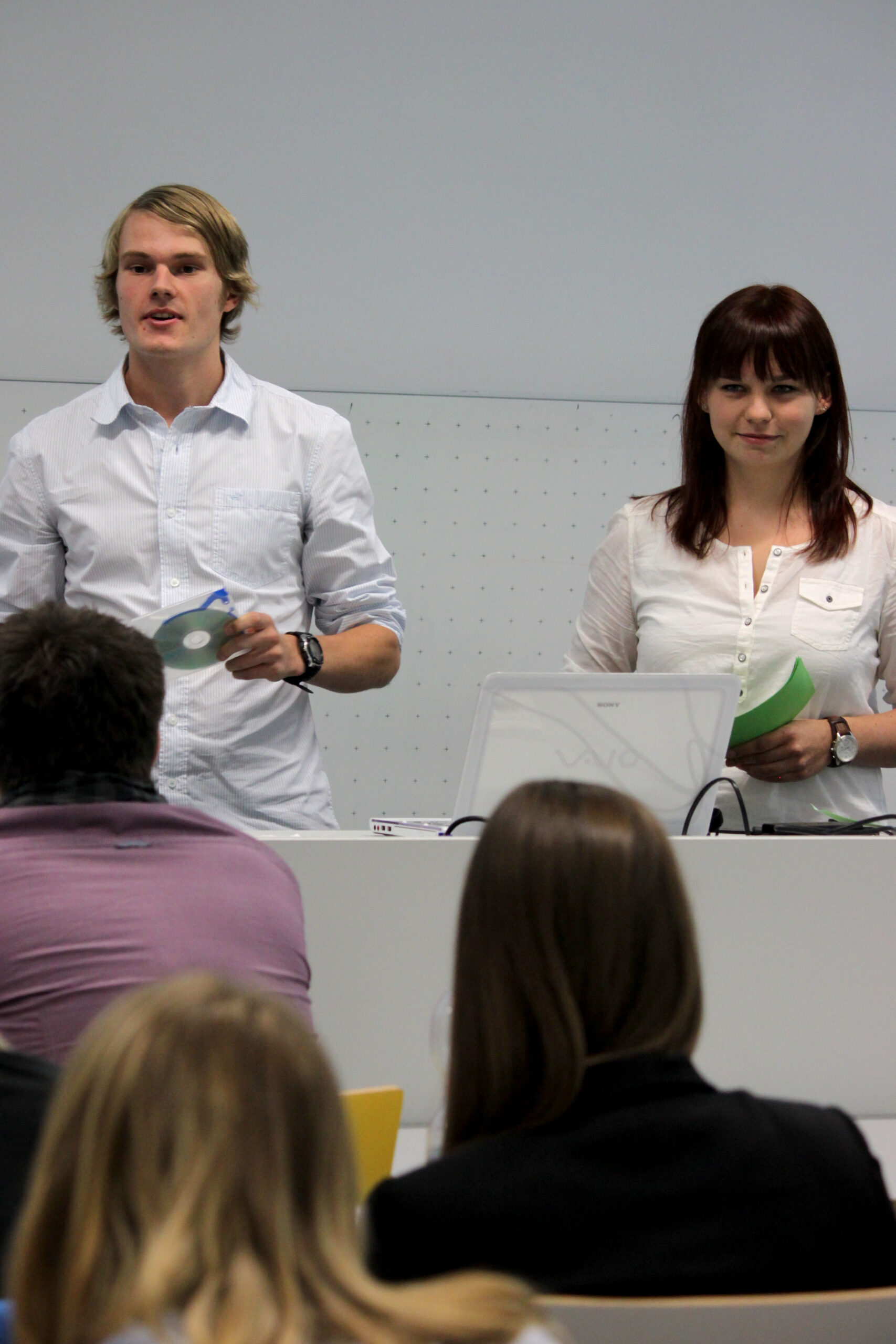 Zwei Personen stehen an einem Podium und halten einen Vortrag vor einem aufmerksamen Publikum an der Hochschule Coburg. Einer hält eine CD in der Hand, während der andere in seinen Notizen nachschlägt. Vor einem schlichten Hintergrund steht ein geöffneter Laptop auf dem Podium und unterstreicht die akademische Atmosphäre dieser spannenden Sitzung.