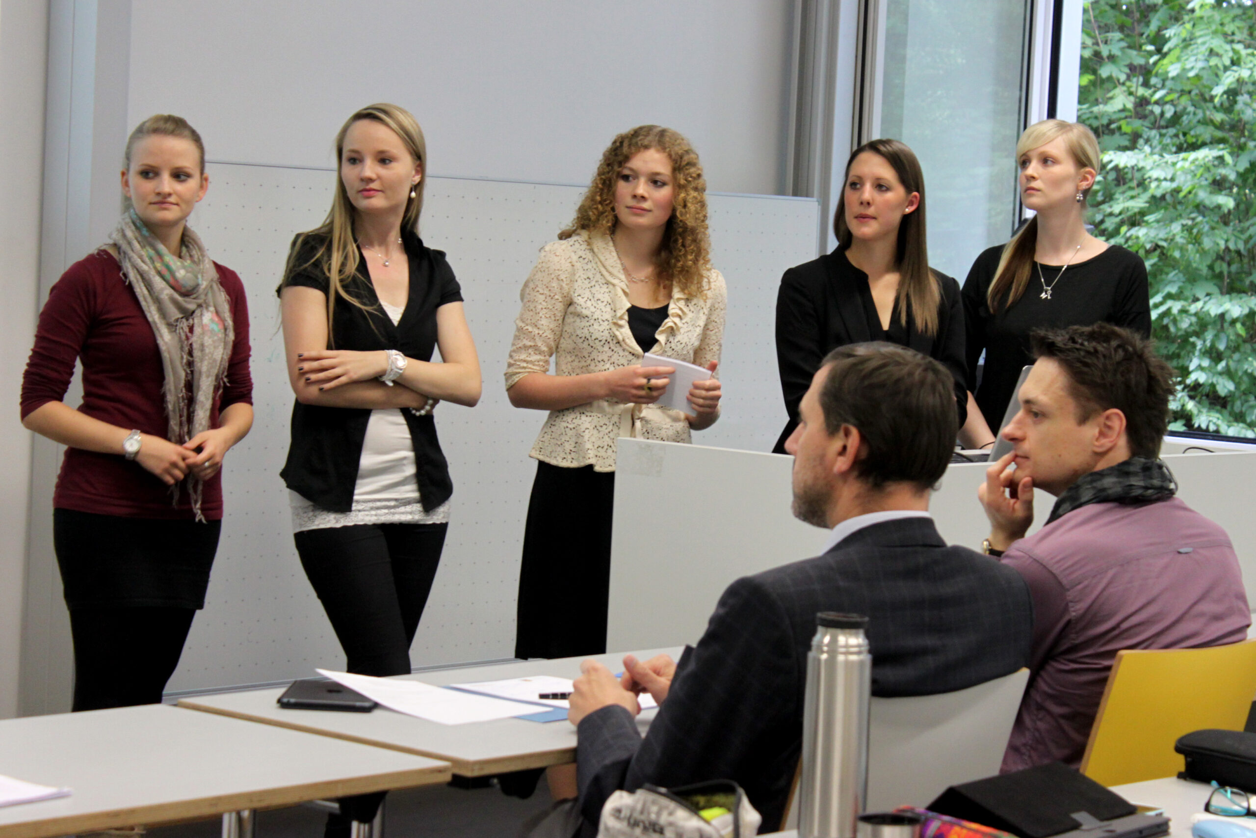 Fünf Frauen, Vertreterinnen der Hochschule Coburg, stehen vor einem Hörsaal und halten einen Vortrag vor drei sitzenden Männern. Die Frauen halten Unterlagen in der Hand und sind leger gekleidet. Der Raum hat große Fenster mit Blick ins Grüne. Die Männer hören aufmerksam zu.