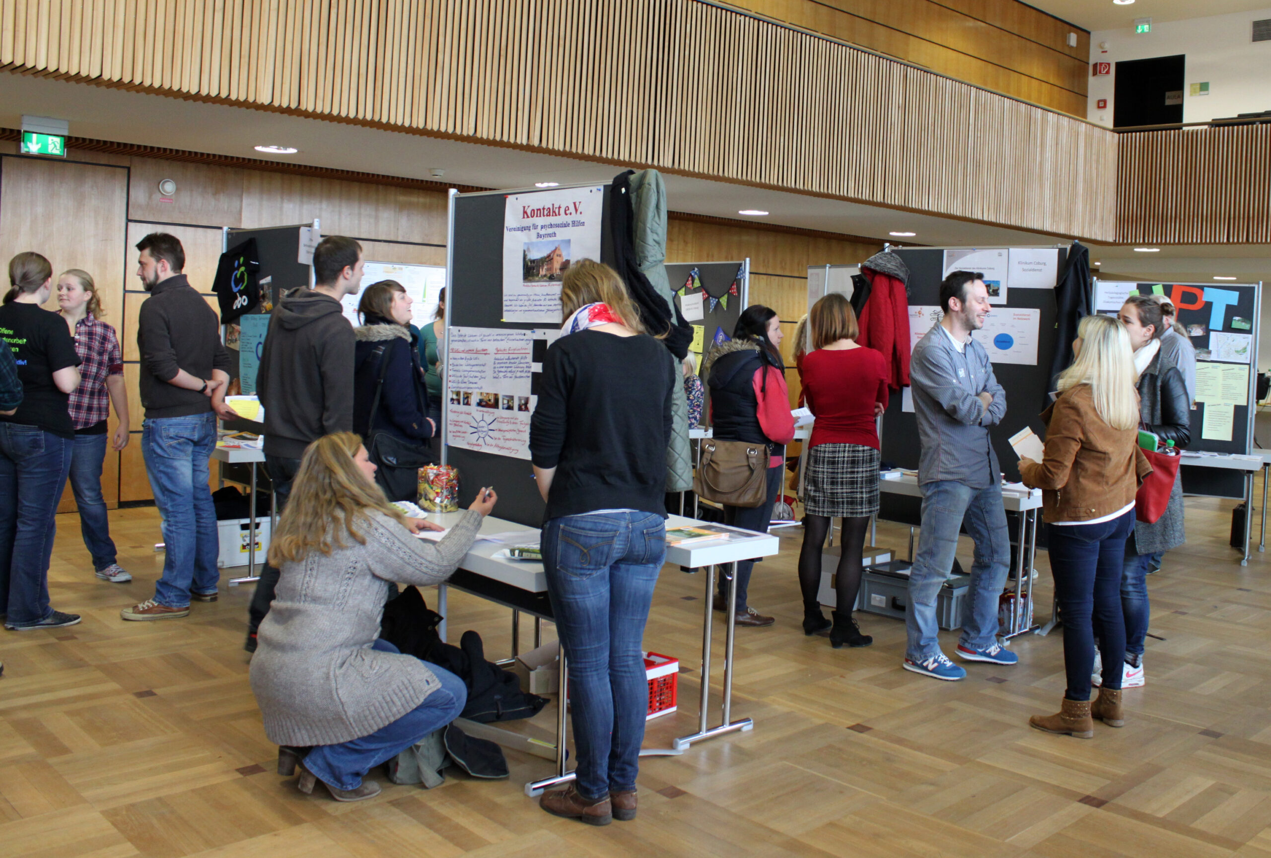 In einem großzügigen Raum mit Holzboden an der Hochschule Coburg haben sich Menschen versammelt, tauschen sich aus und betrachten mehrere Schautafeln, die auf Tischen aufgestellt sind. Einige lesen Plakate, andere unterhalten sich. Die Atmosphäre wirkt locker und lebhaft.