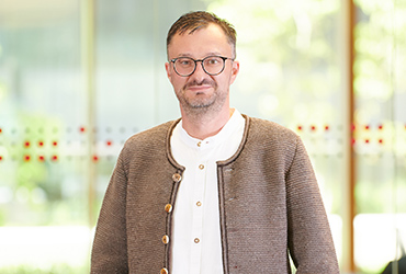 Ein Mann mit Brille und einer braunen Strickjacke über einem weißen Hemd steht in einem Innenbereich, möglicherweise in den Fluren der Hochschule Coburg, vor unscharfem Hintergrund.