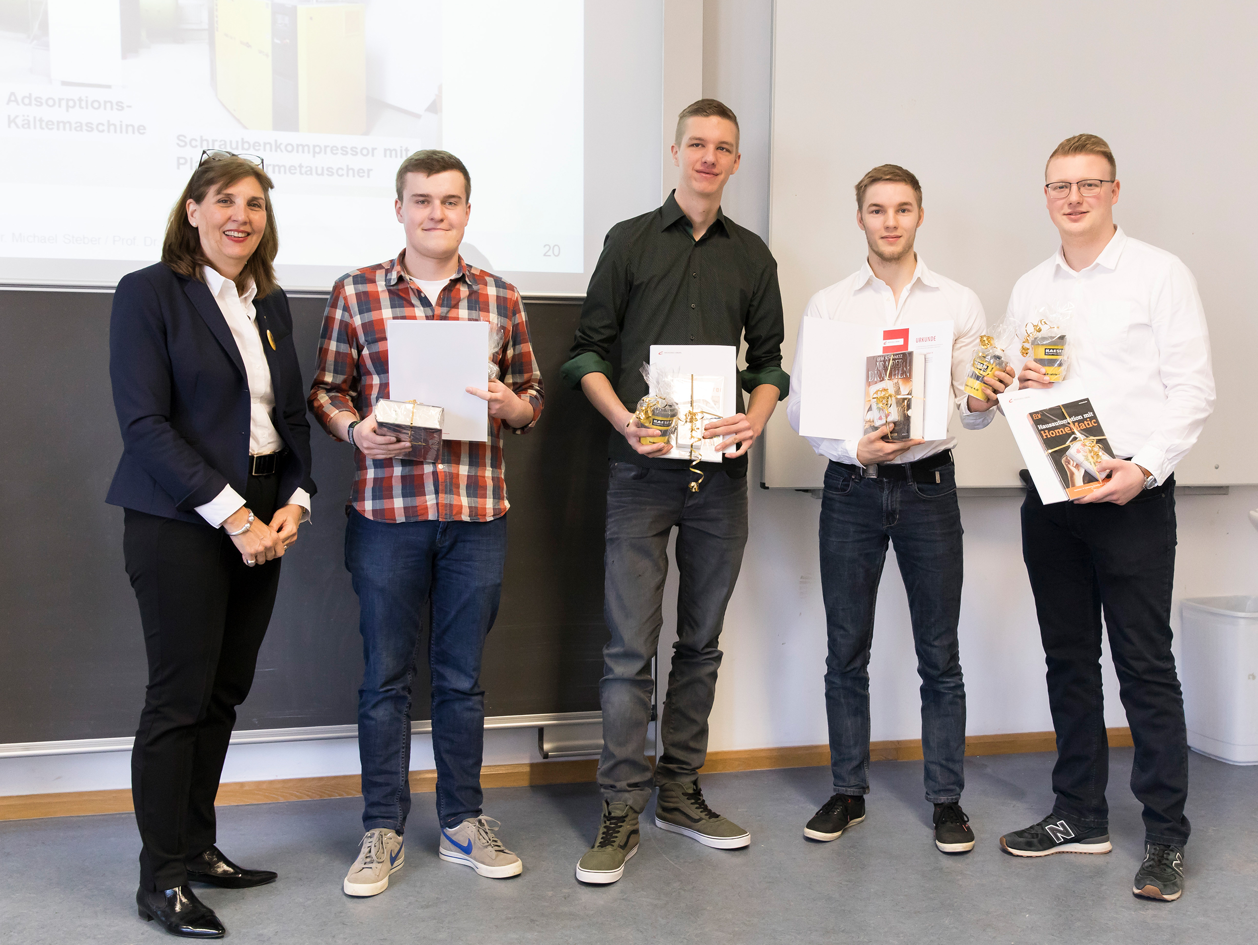 Eine Gruppe von fünf Personen steht zusammen in einem Hörsaal der Hochschule Coburg, hinter ihnen eine Tafel. Vier von ihnen halten Zertifikate und kleine Geschenke in den Händen. Eine Person links ist eine Frau in Berufskleidung, die der Gruppe herzlich zulächelt.