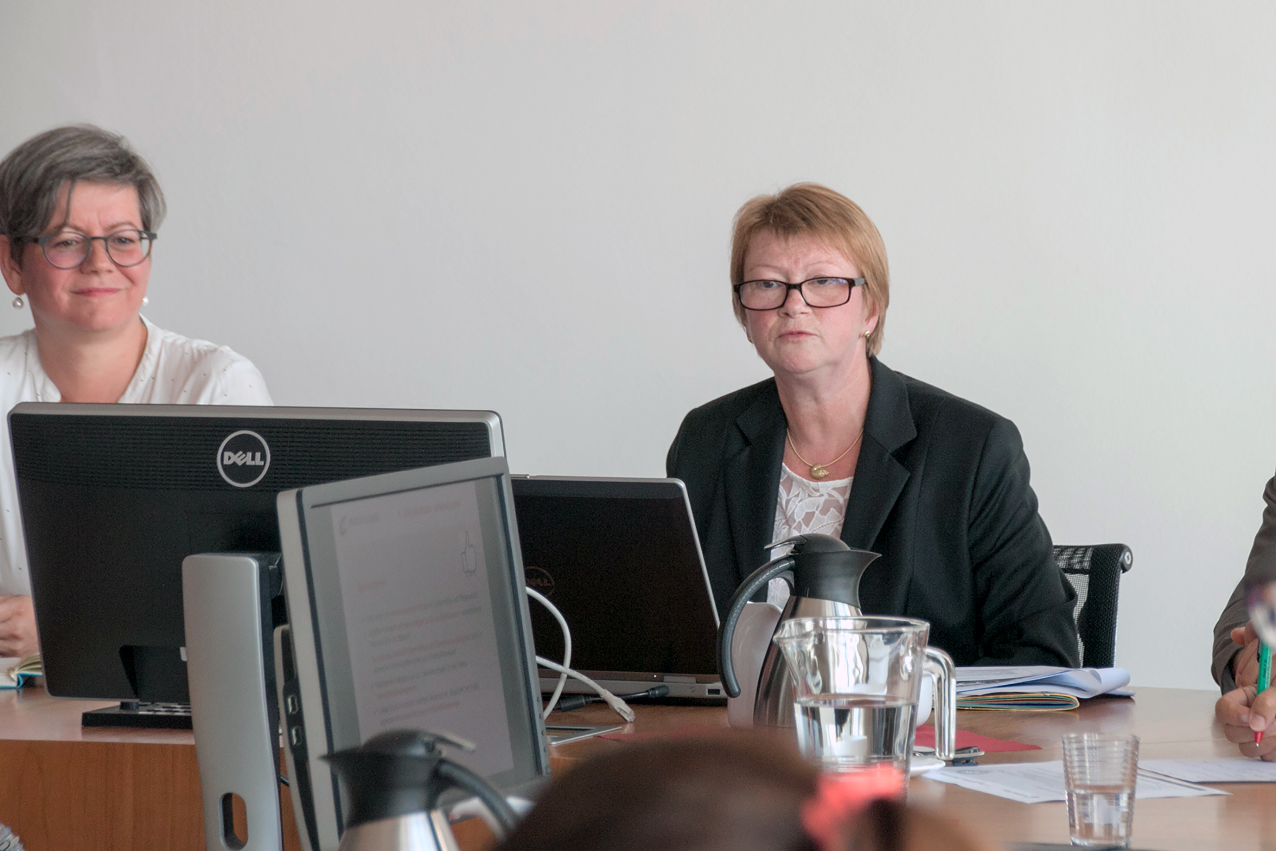 Zwei Frauen sitzen mit Laptops und Dokumenten an einem Schreibtisch und verkörpern damit die professionelle Atmosphäre der Hochschule Coburg. Eine trägt einen schwarzen Blazer und eine Brille, die andere ein weißes Hemd. Das Meeting ist mit Wasserkrügen gedeckt, alles vor einem neutralen Hintergrund, der Konzentration und Zusammenarbeit betont.